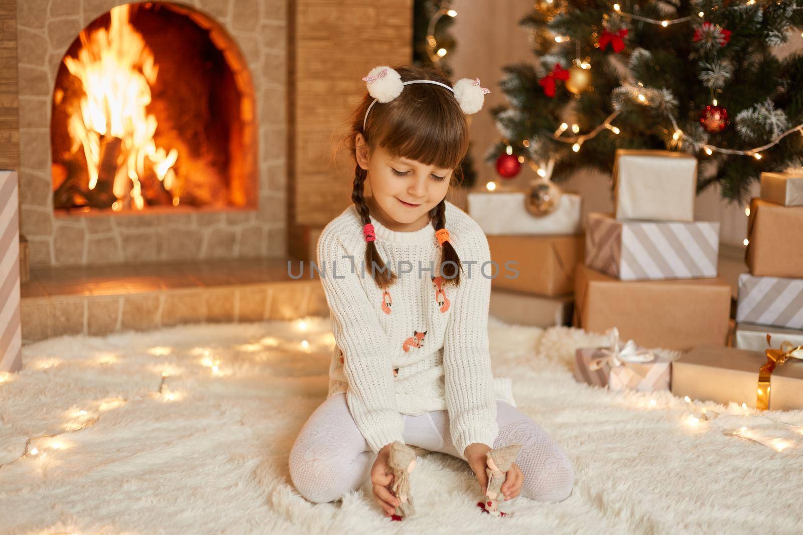 Little girl near christmas tree and fireplace playing with tiny toys, her x-mas present, child with pigtails sitting on floor on carpet, wearing white pullover.