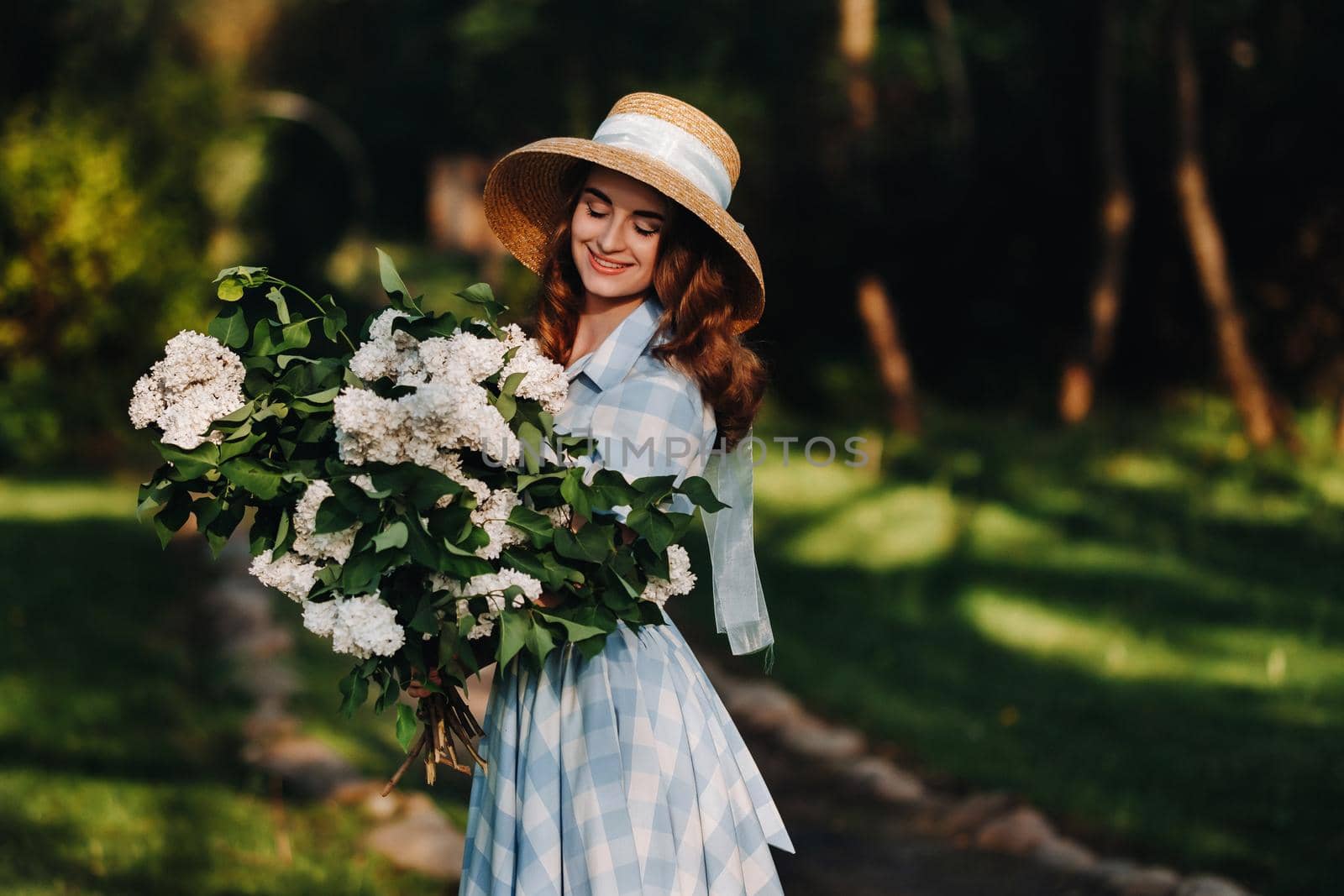 Portrait of a beautiful girl with long hair, a straw hat and a long summer dress with lilac flowers in the garden.