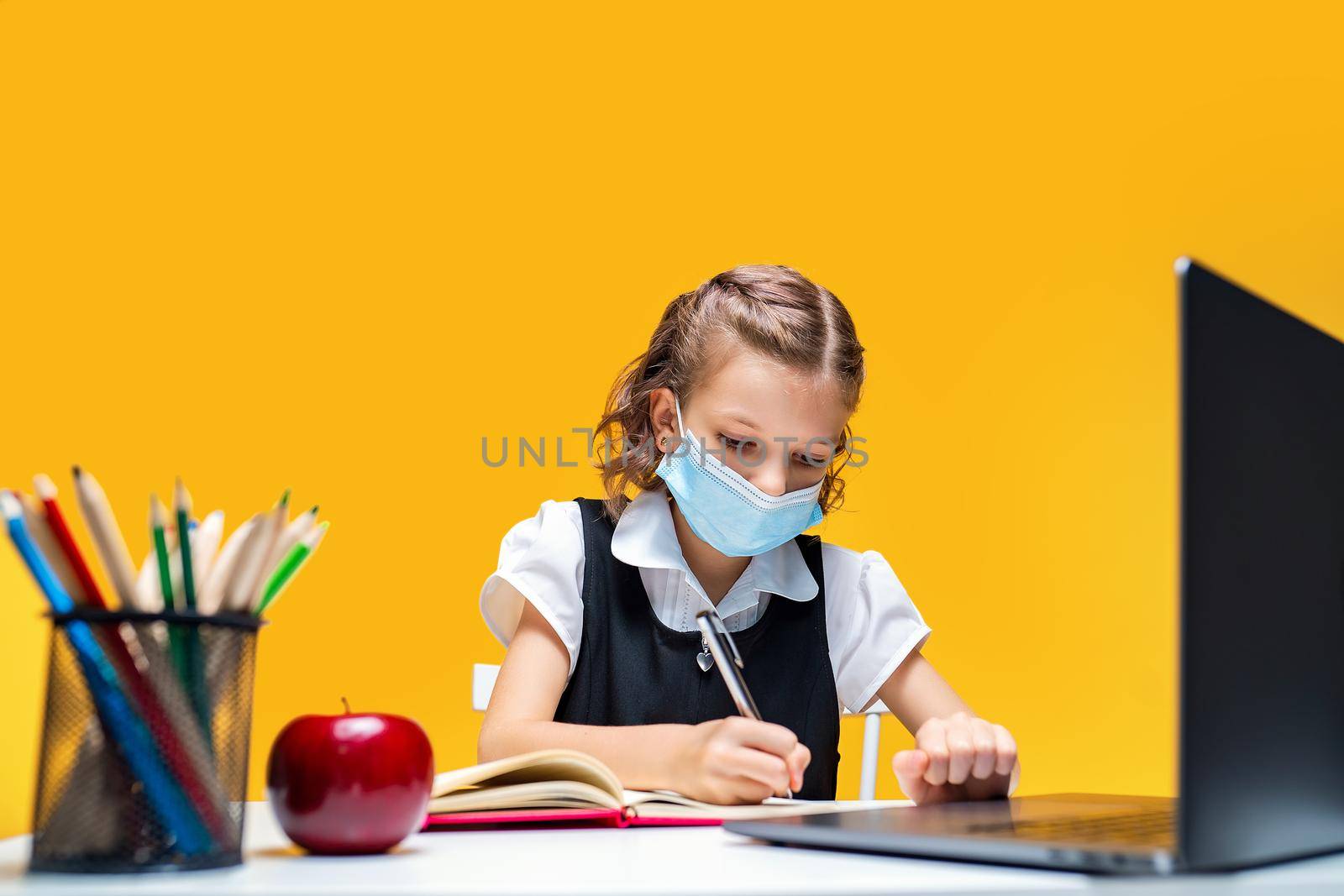 Serious schoolgirl in mask doing her homework with laptop. Writing in notebook. Distance education by Studio_SOK
