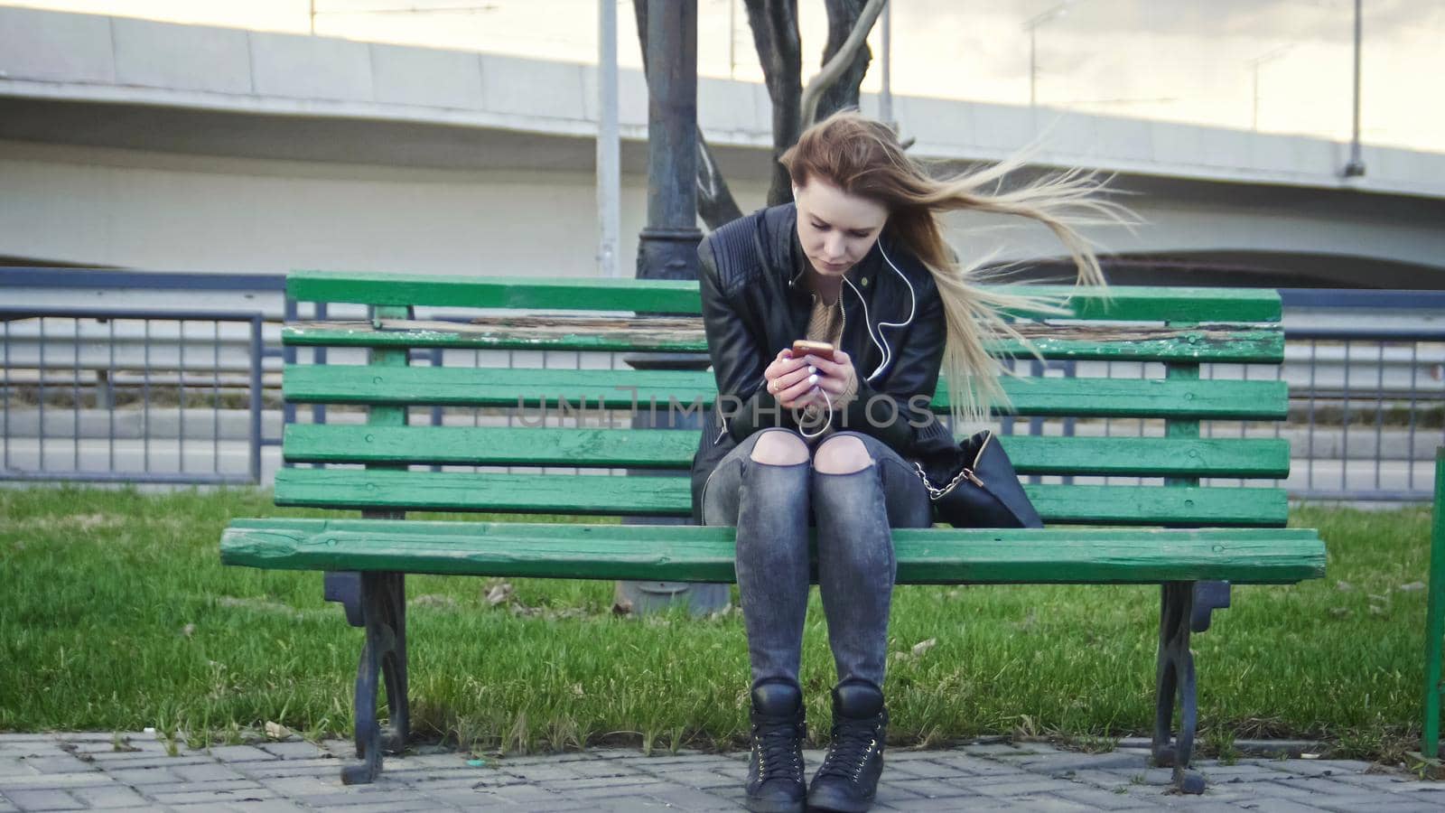 Dissapointed girl with long blonde hair in leather jacket straightens hair use gadget sitting on the bench in the wind listen headphones, wide angle, front view, telephoto