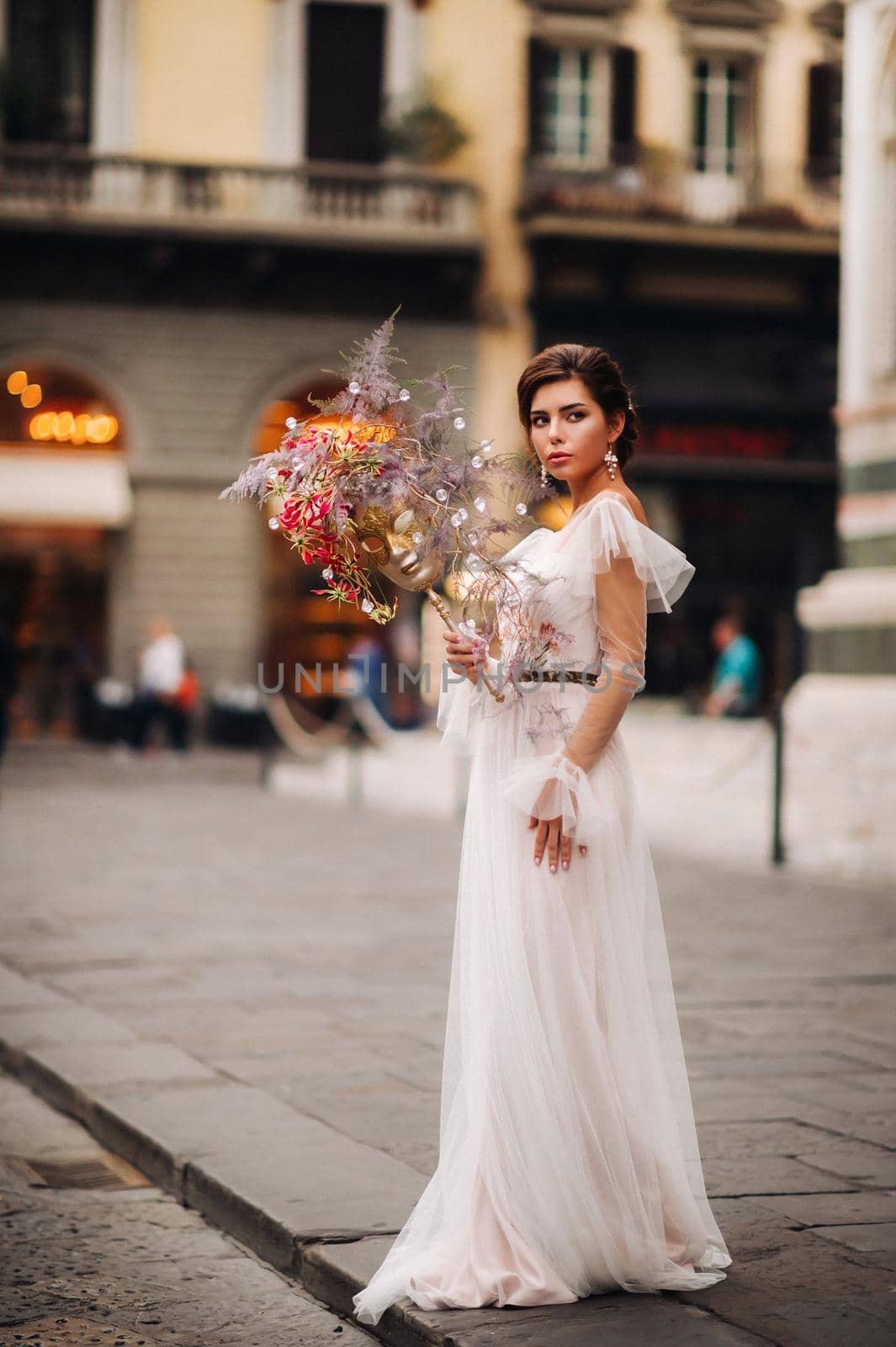 the girl-bride is with beautiful flower pattern as a mask in Florence, stylish bride in a wedding dress standing with a mask in the Old town of Florence. Model girl in Florence.