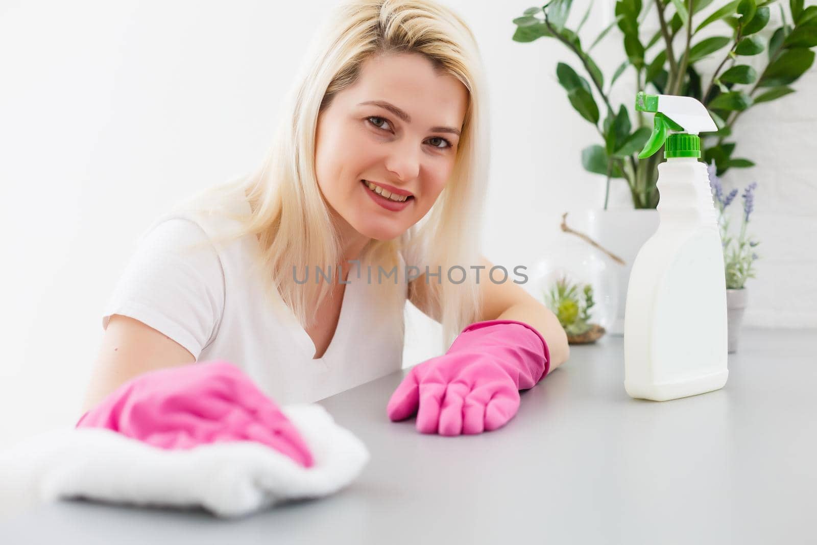 Woman in the kitchen is smiling and wiping dust using a spray and a duster while cleaning her house, close-up