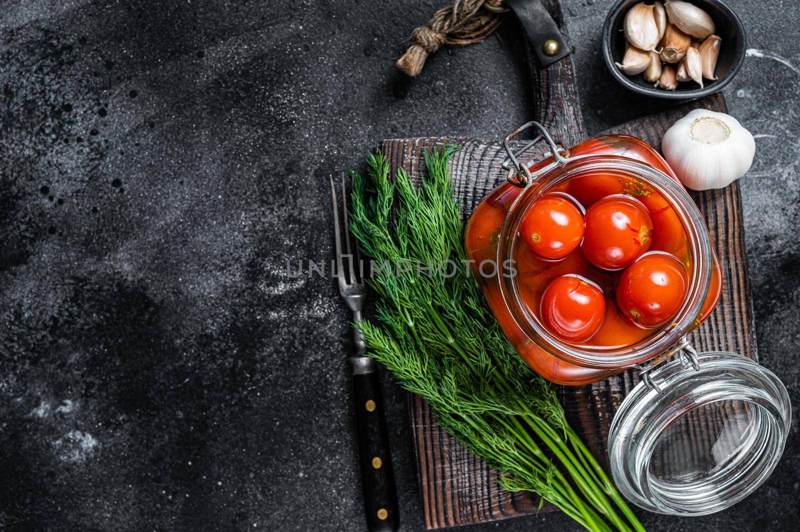 Pickled cherry tomatoes in a glass jar with herbs. Black background. Top view. Copy space.