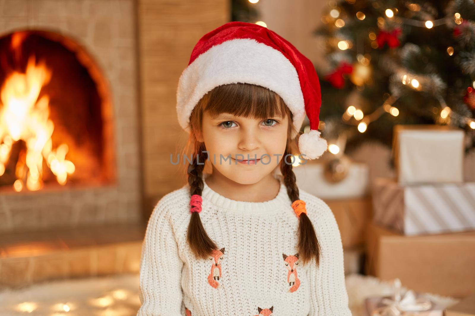 Happy smiling child girl posing at home in decorated living room, kid looking directly at camera, has pigtails, wearing white jumper and red santa claus hat, posing near christmas tree and fireplace.