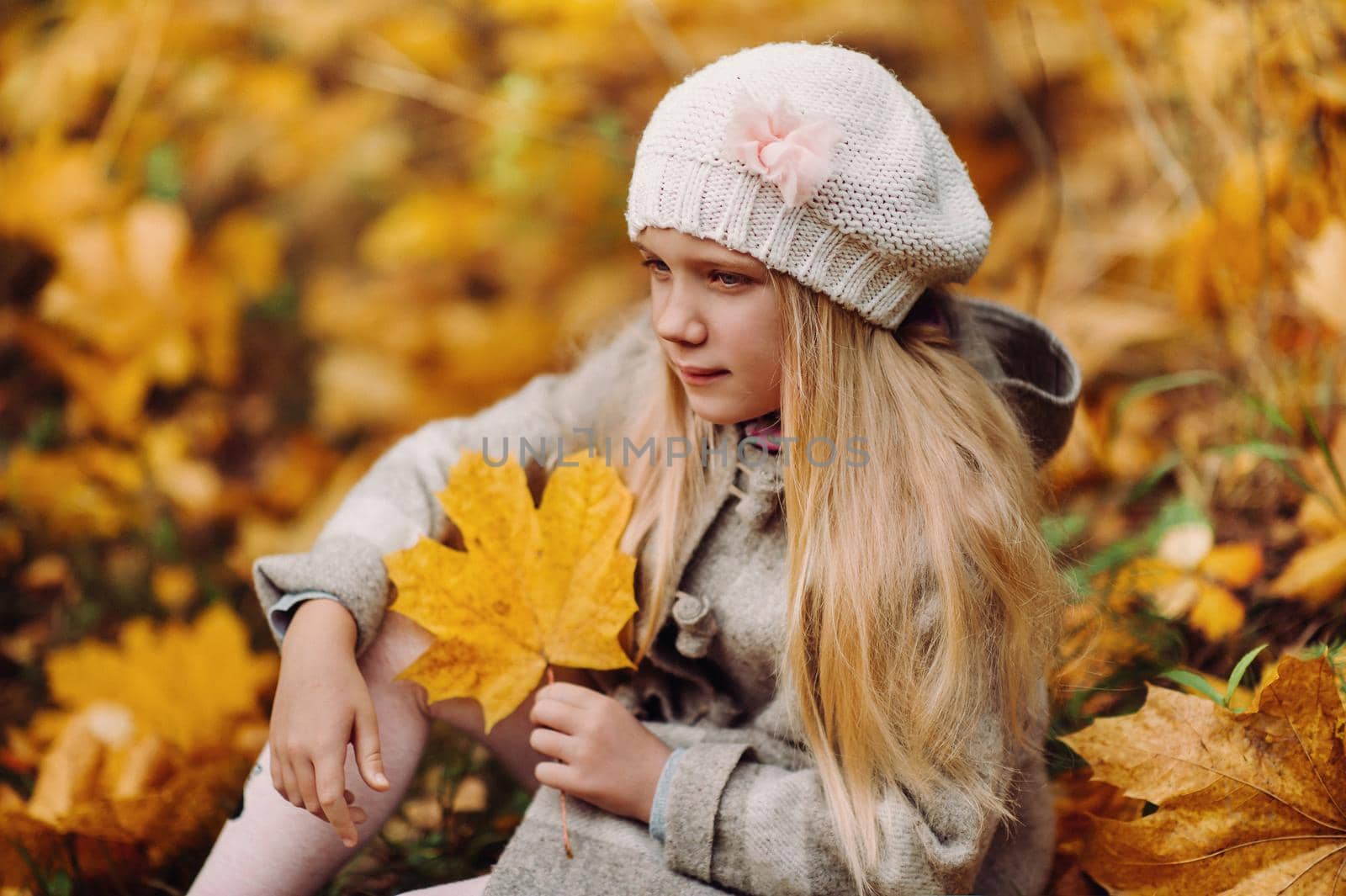 A charming little girl in a coat sits in autumn with a leaf in her hands.