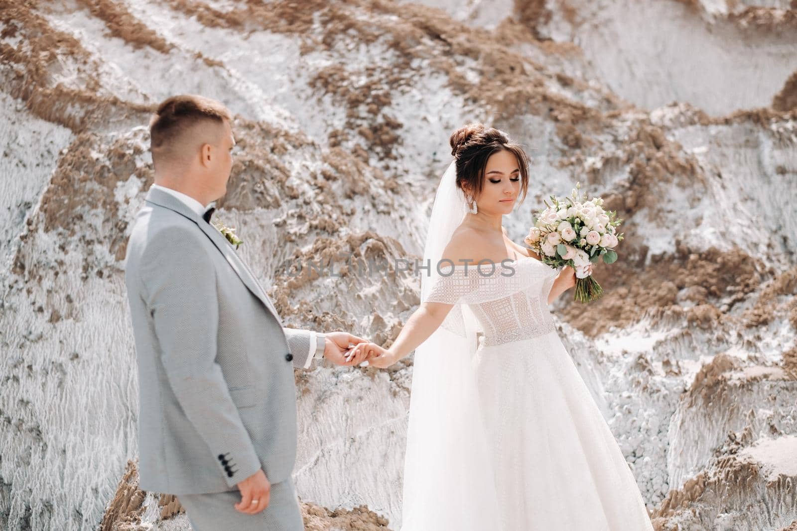 A beautiful couple of lovers posing in a white salt mountain. A young woman in a stylish wedding dress and a beautiful stylish man in a gray suit. The concept of the wedding day. by Lobachad