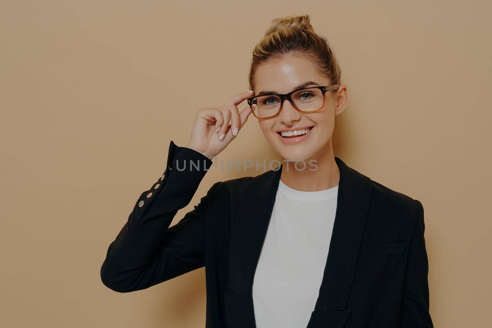 Positive happy young woman touching rim of glasses and looking through them, smiling female student touching spectacles while looking at camera with smile, isolated over beige wall with copy space