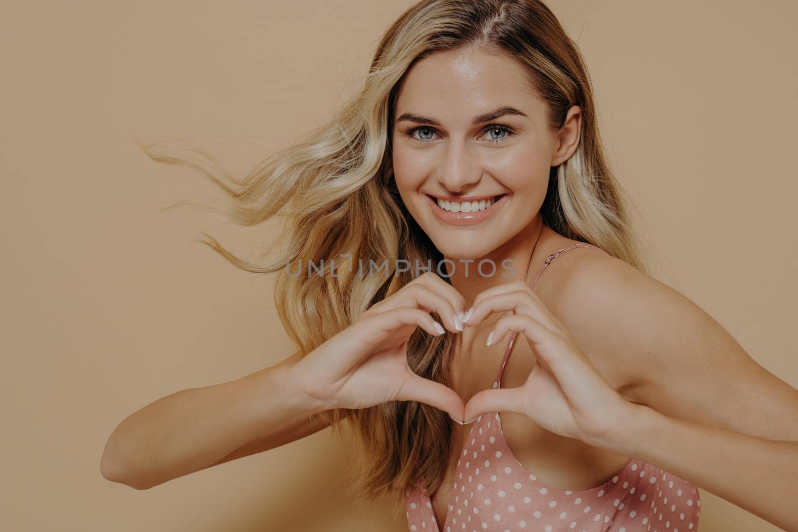 Upbeat blonde young woman in pink dress showing heart gesture with hands, displaying her love and affection with happy tender smile, standing isolated next to orange background