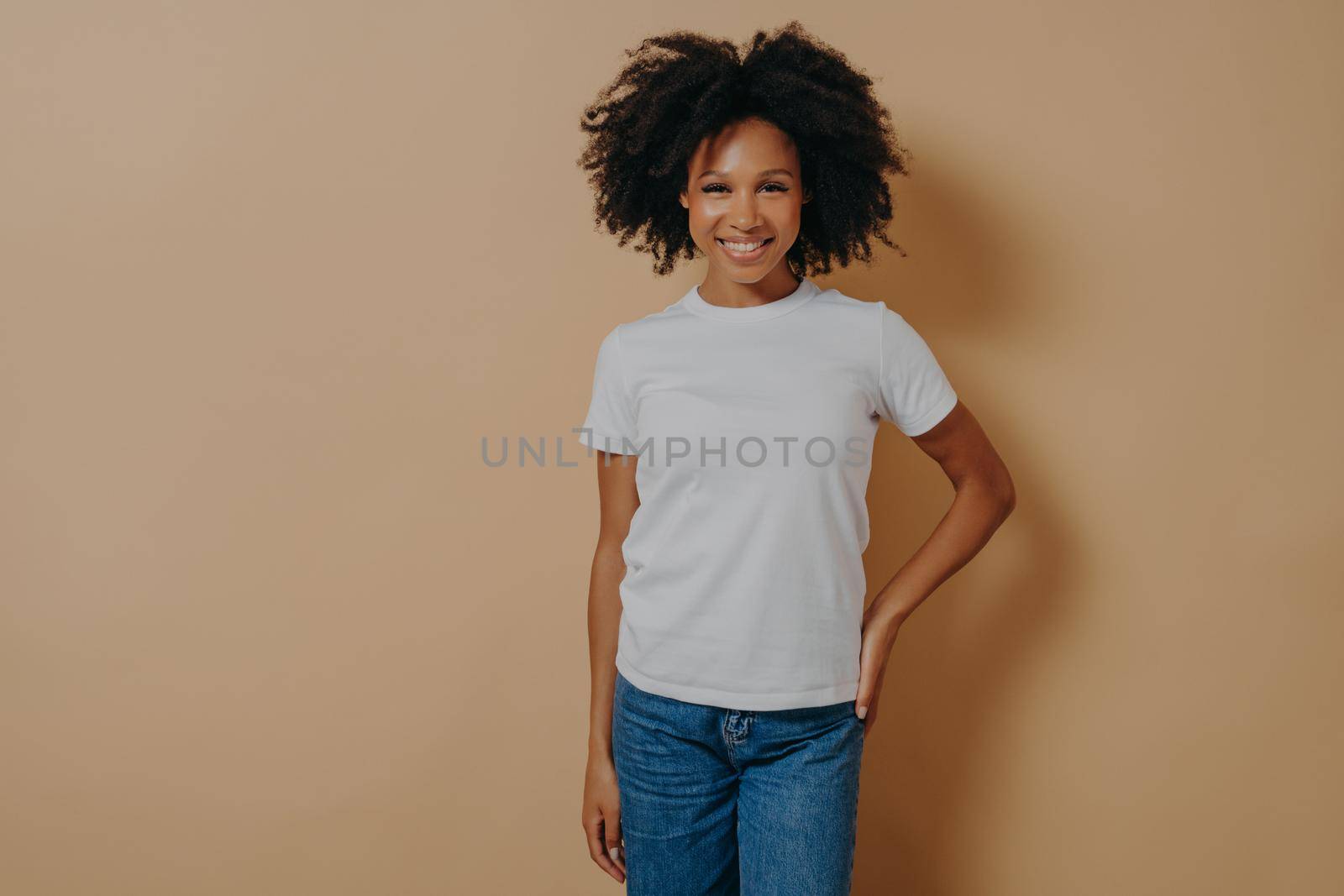 Beautiful cheerful dark skinned woman in casual outfit smiling at camera, cheerful african female in white tshirt and jeans expressing positive emotions, posing against beige background in studio