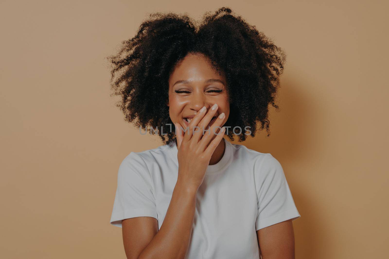 Close up studio portrait of happy biracial girl. Beautiful dark skinned millennial woman covering her mouth with hand while laughing. Cute positive mixed race female isolated over beige background