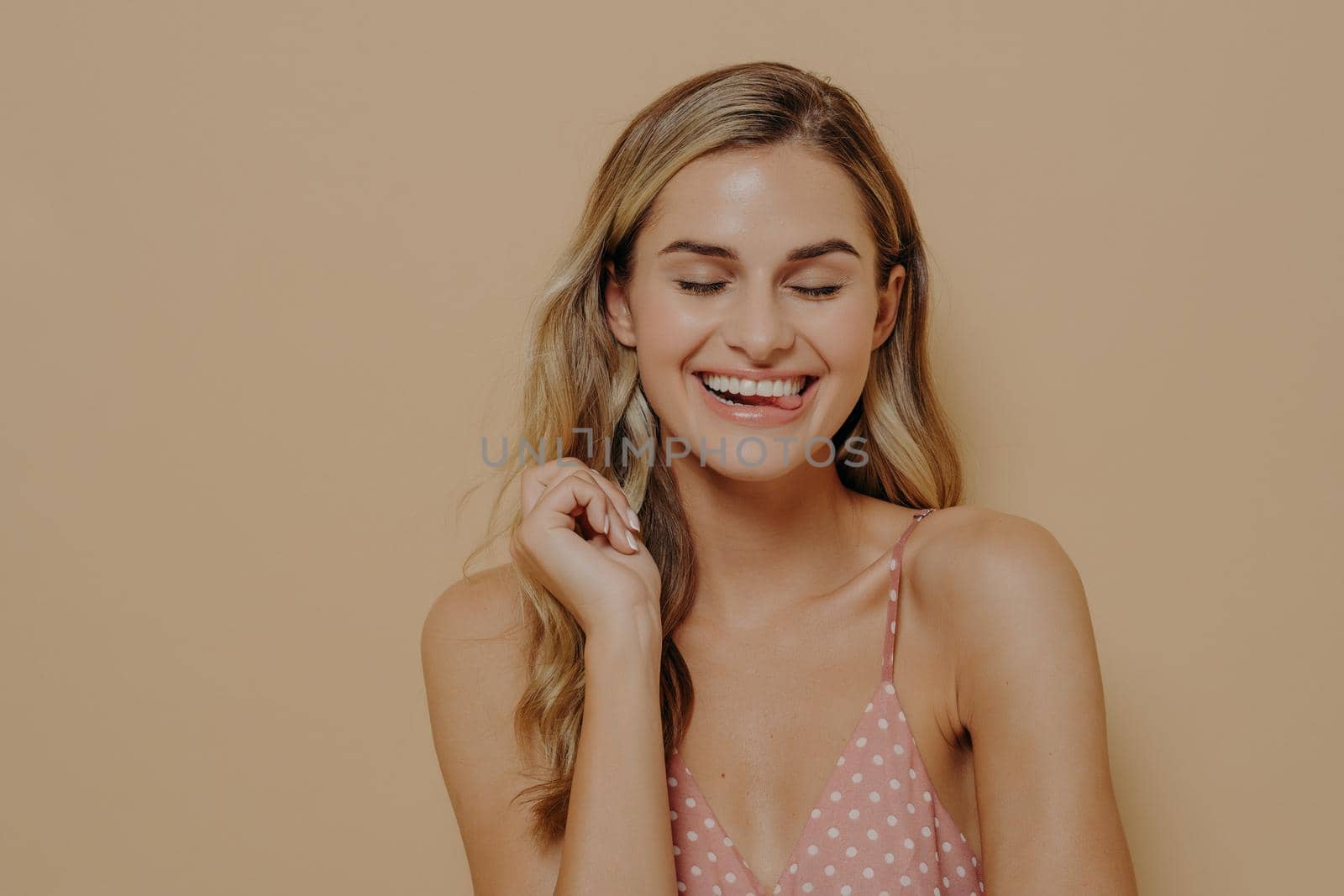 Human face expressions. Joyful cheerful young beautiful woman with fair wavy hair in polka dot pink summer dress laughing at joke, smiling broadly with closed eyes. Positive emotions concept