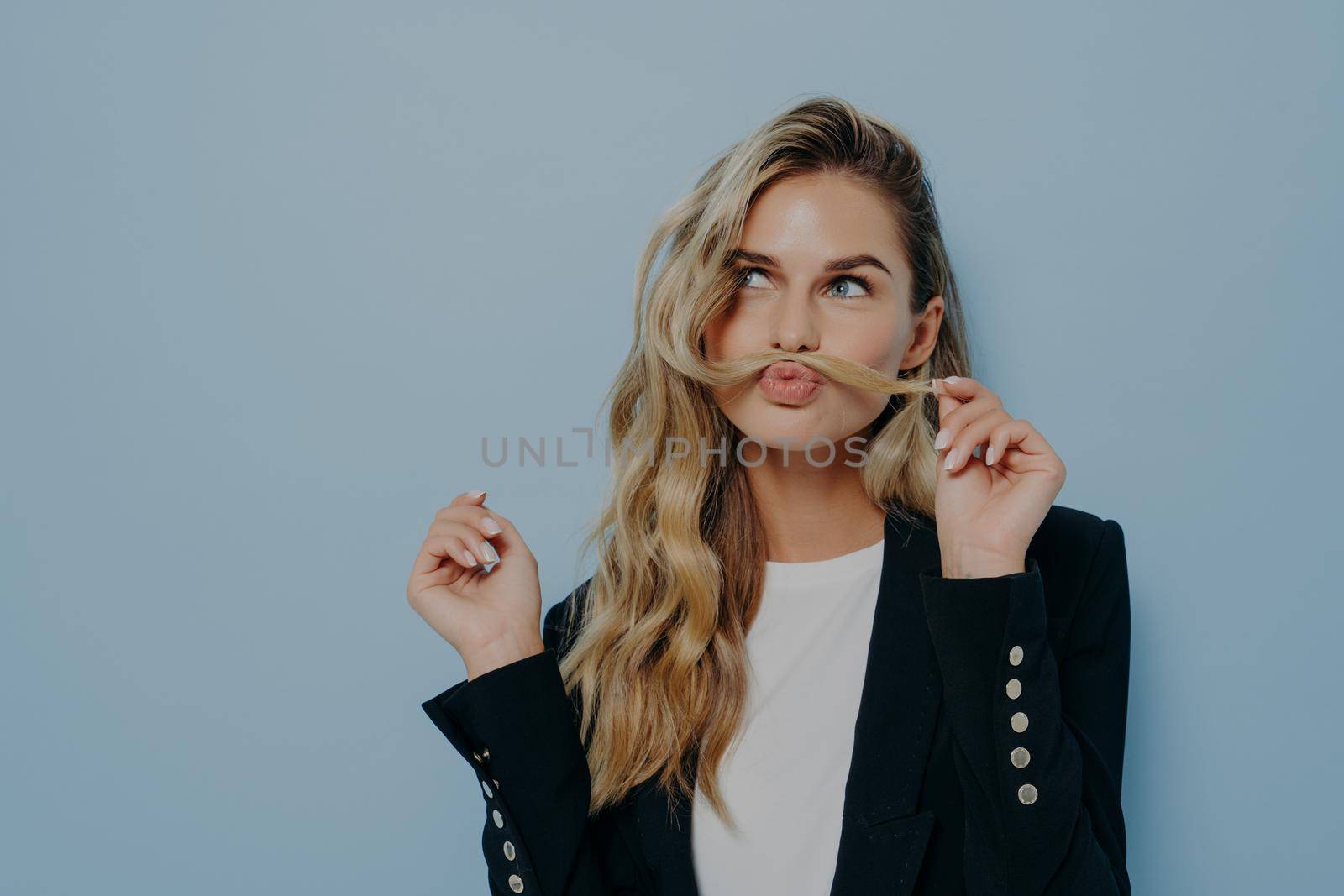 Funny joyful blonde woman having fun while posing against blue studio wall, playing with strand of hair by vkstock