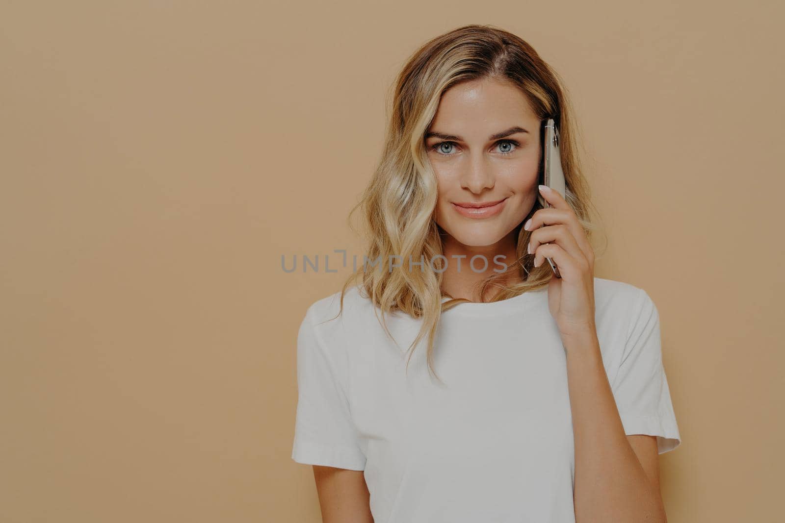 Confident female with blonde dyed hair holding smartphone and calling to friend, looking at camera with smirk while posing in studio, dressed in white t shirt. Modern technologies and communication