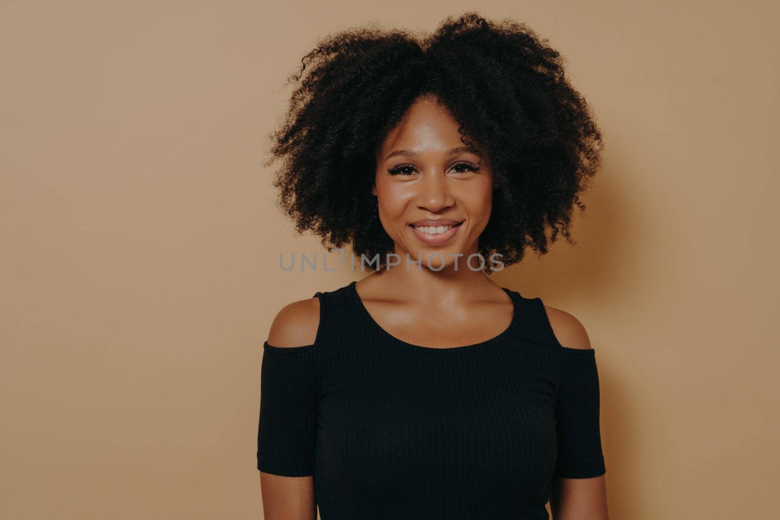 Portrait of cheerful smiling beautiful dark skinned girl in black tshirt standing isolated against dark beige wall background with copy space for advertising. Youth and happiness concept