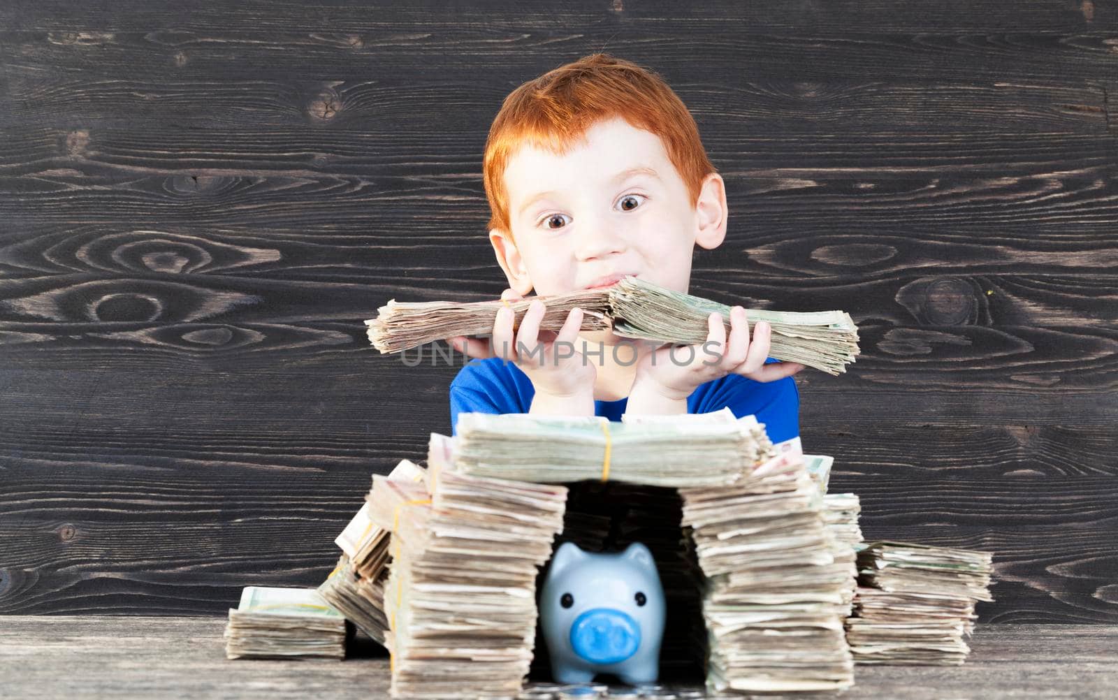 red-haired boy built for his blue pig piggy bank with coins house of packs of paper money, close-up of a child holding a wrapped bill