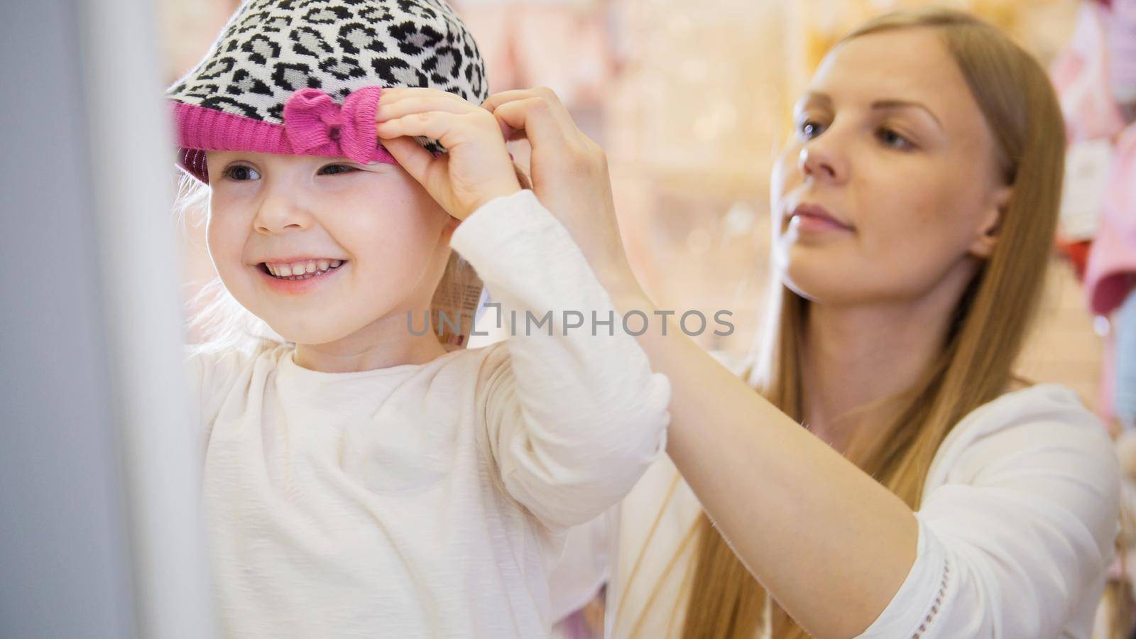 Kids dress store - little blonde baby girl with mother doing shopping and buying red hat, telephoto