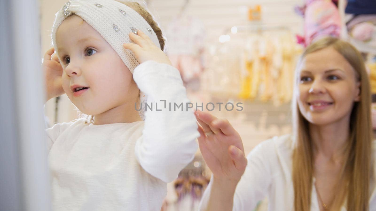 Baby's dress store - little blonde baby girl with mother doing shopping and buying pretty hat, telephoto