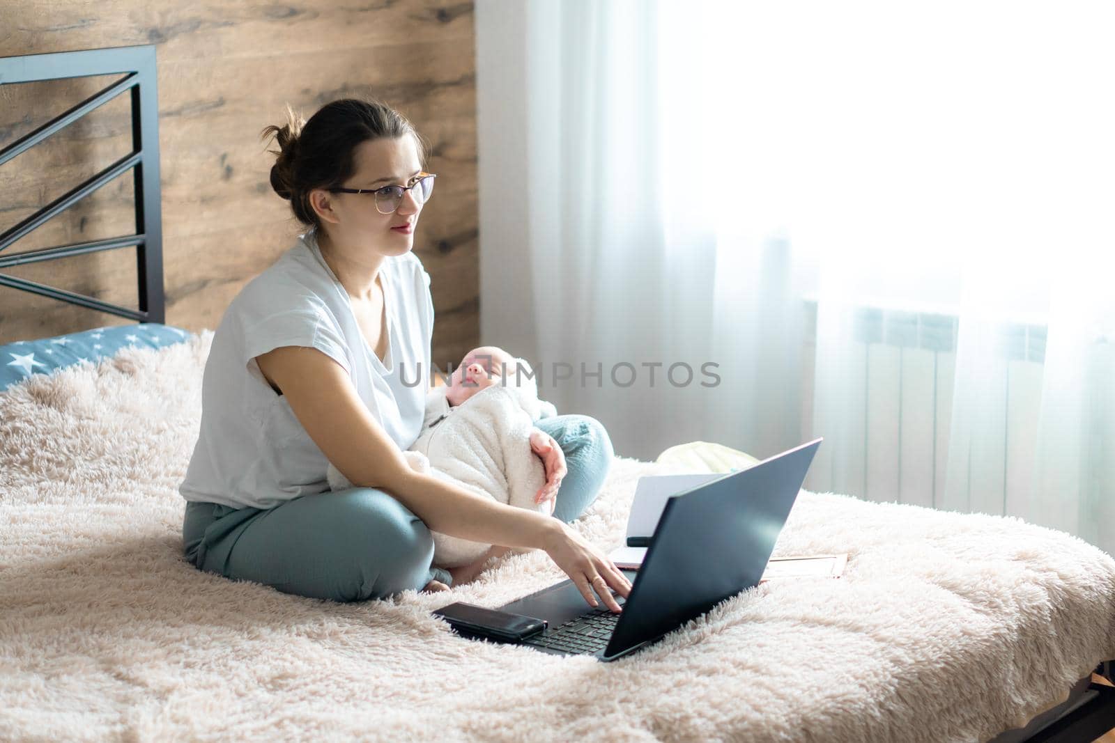 remote work, training, work at home, femininity, technology, leisure, communication online concept - Young cute girl with newborn baby sits on bed with laptop and works remotely, copy space