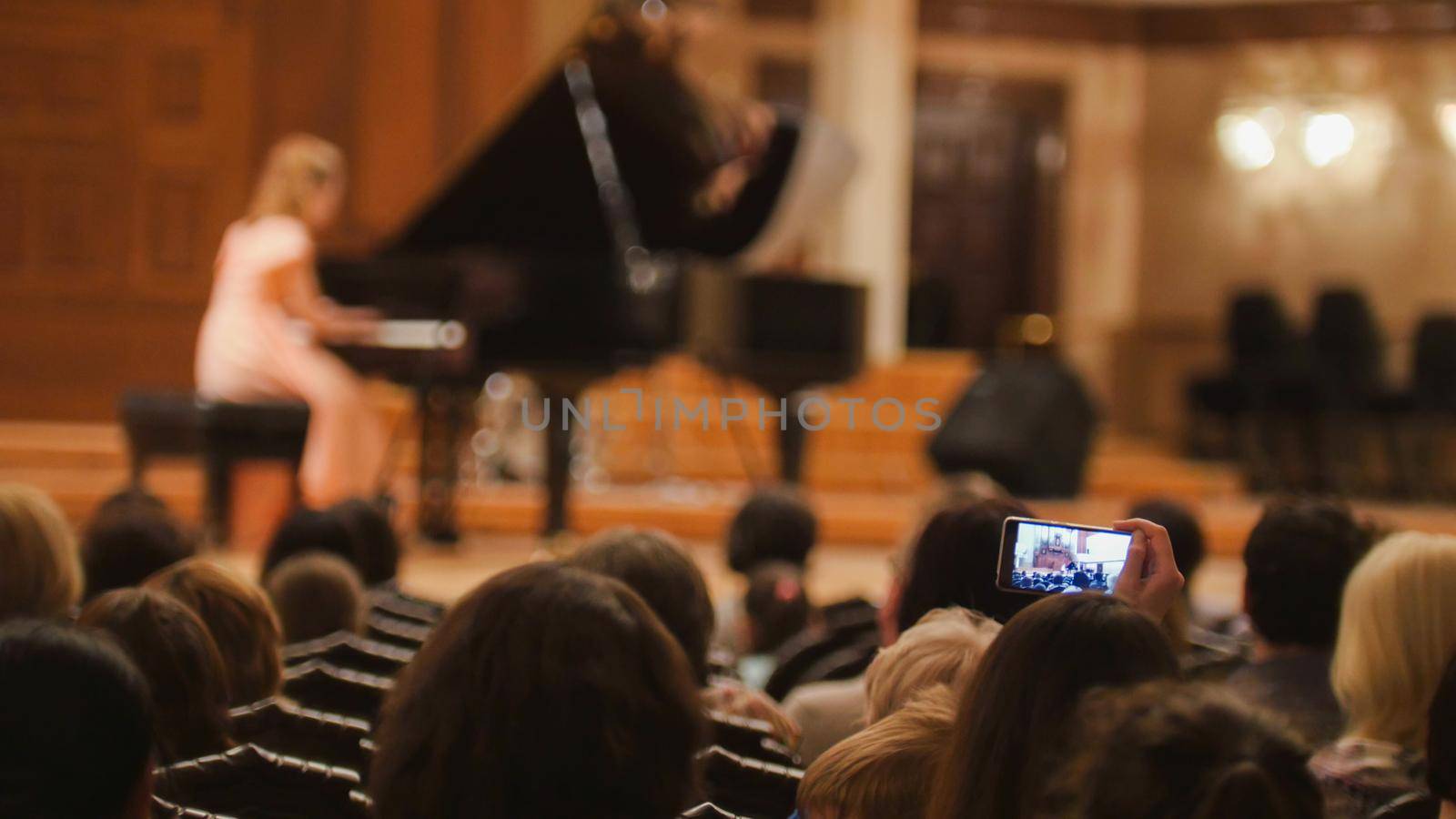 Spectators in concert hall during performing piano girl- people shooting performance on smartphone, telephoto