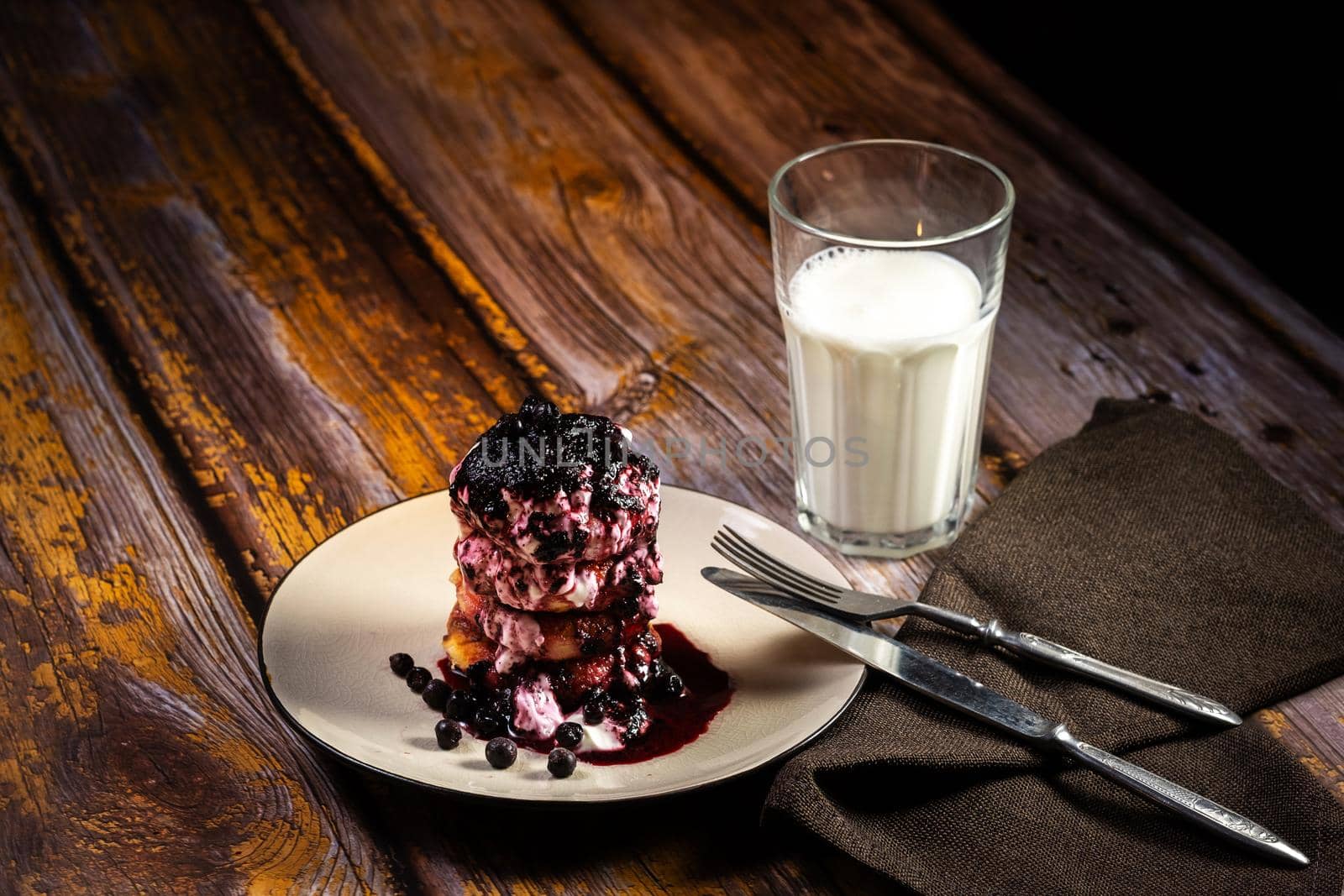 Fried cheesecakes with blueberry jam and sour cream on a plate and a glass of milk on the table by Lobachad
