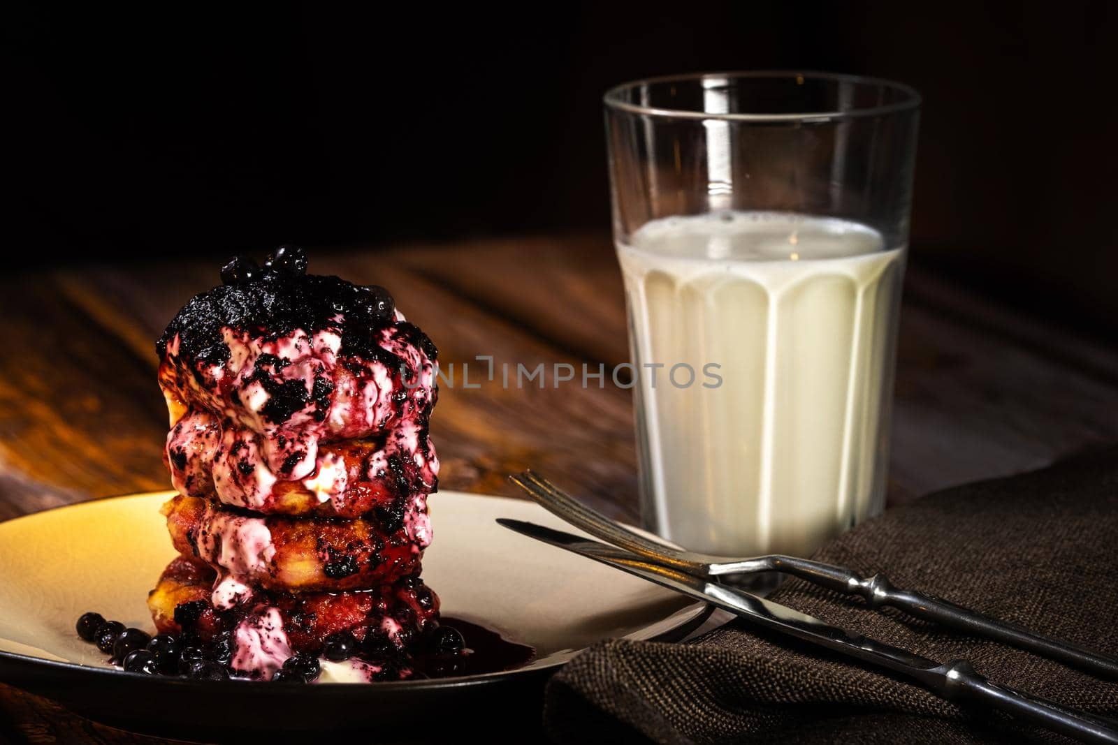 Fried cheesecakes with blueberry jam and sour cream on a plate and a glass of milk on the table.