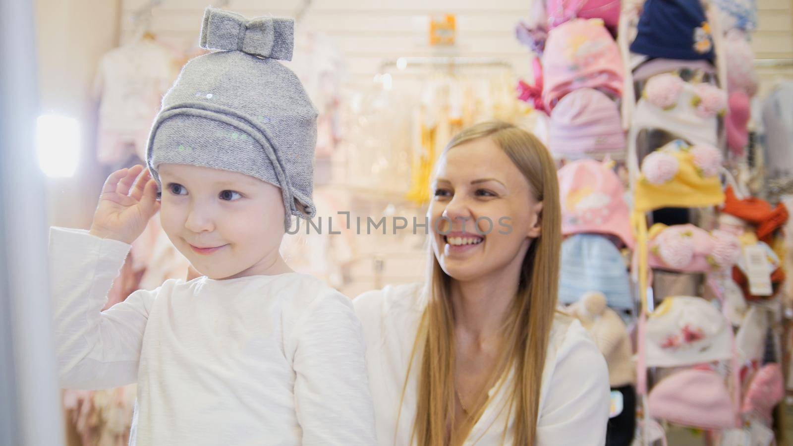 Kids dress store - little blonde baby girl with mother doing shopping and buying hat, telephoto