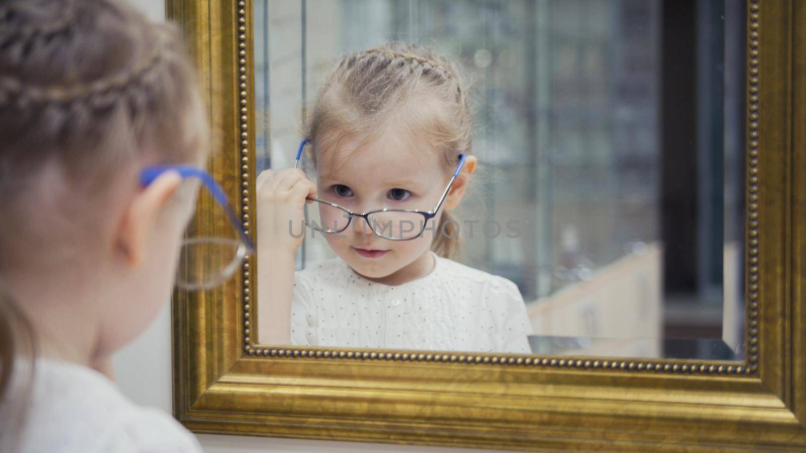 Little girl tries new glasses near mirror - shopping in ophthalmology clinic, close up