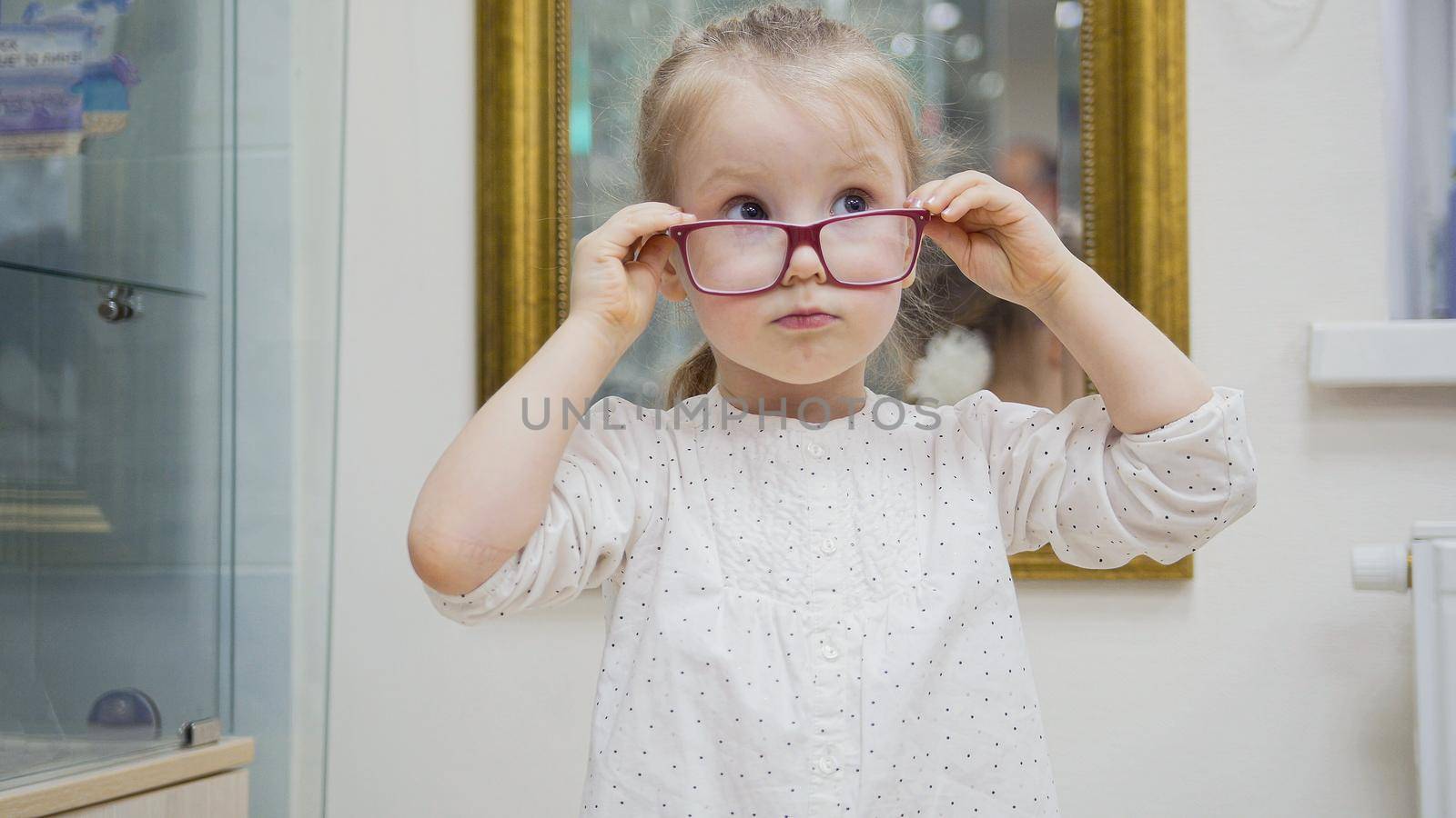 Little girl tries fashion medical glasses near mirror - shopping in ophthalmology clinic, close up