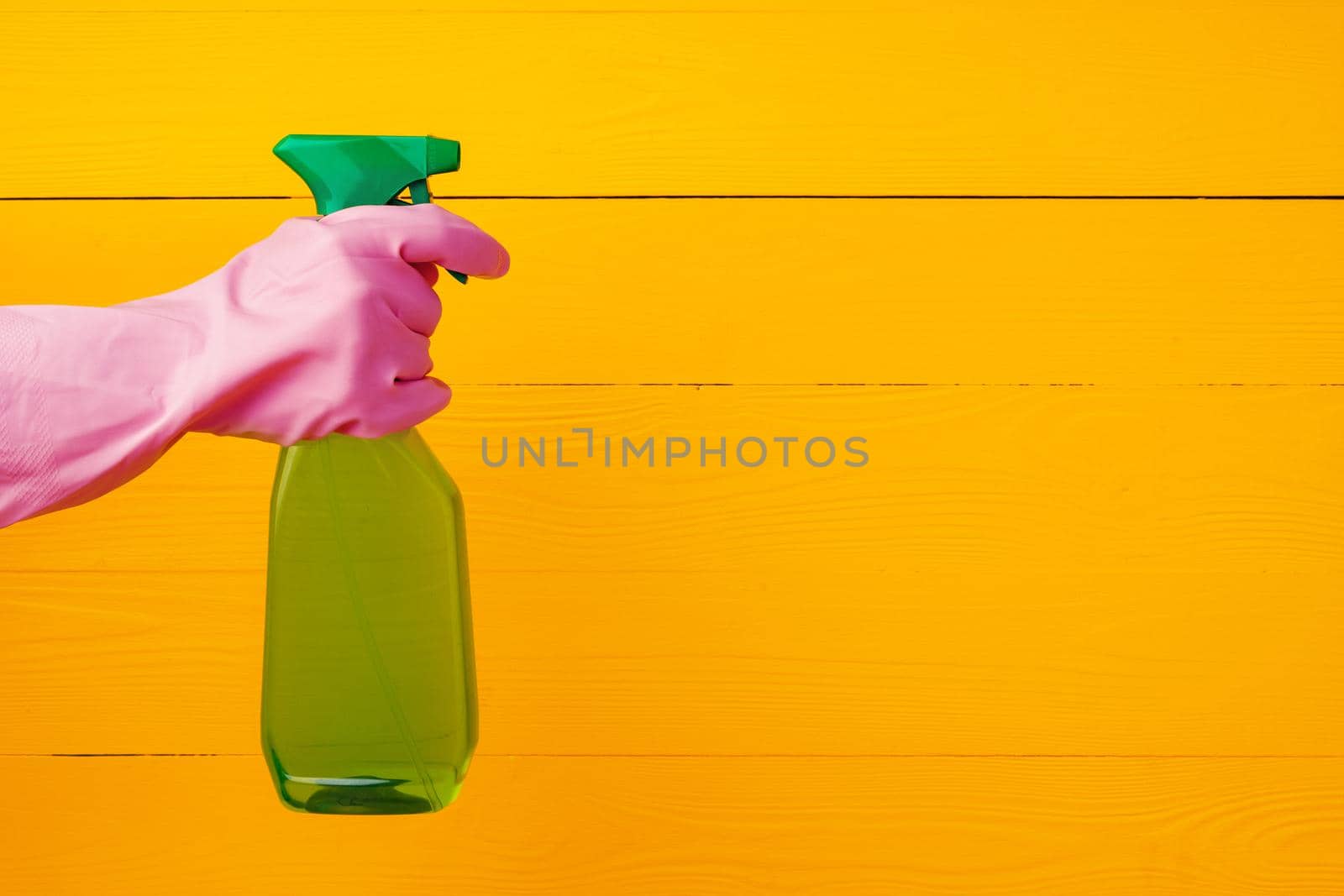 Hand in glove holding household detergent spray against yellow wooden background, close up