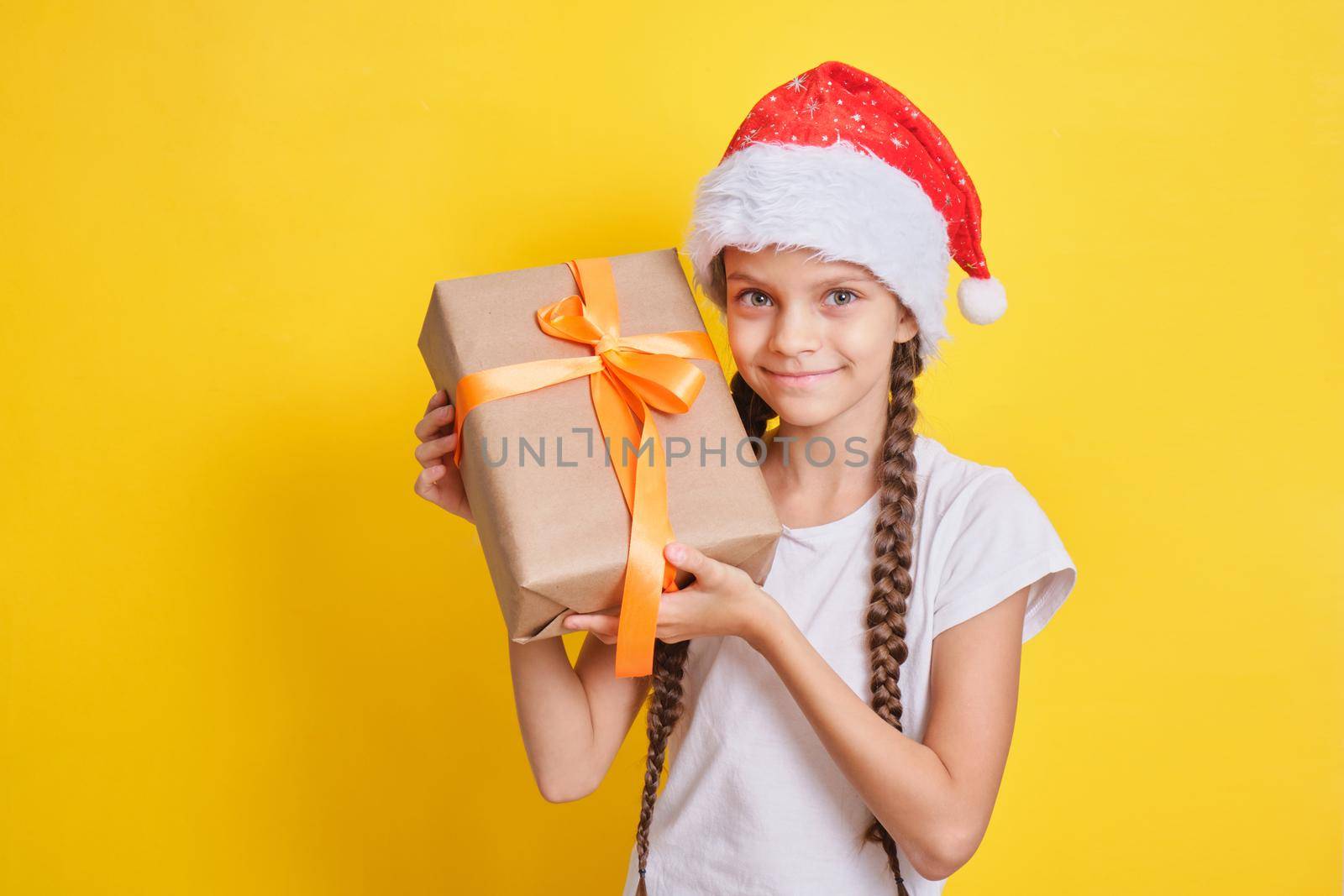 emotional teenager girl in santa claus hat with a gift on a yellow background, christmas gifts concept by natashko