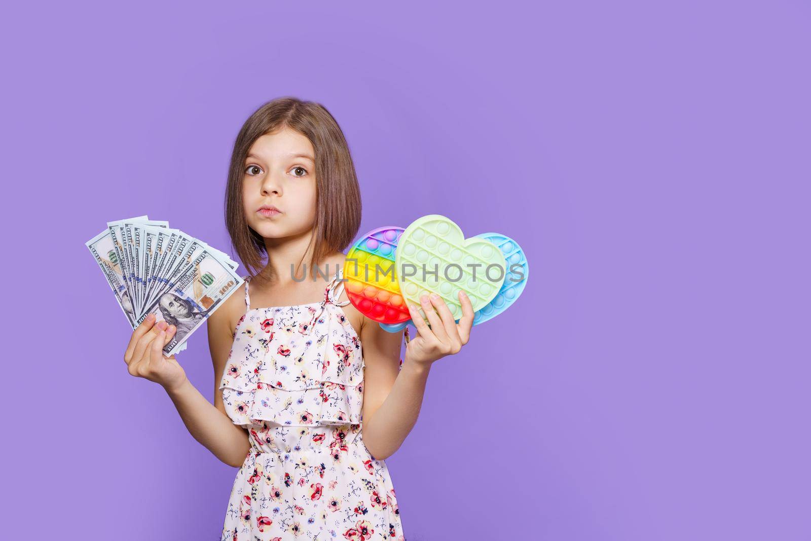 A little girl in a summer dress holds pop it in one hand, US dollar cash in the other and looking at camera. Purple background studio