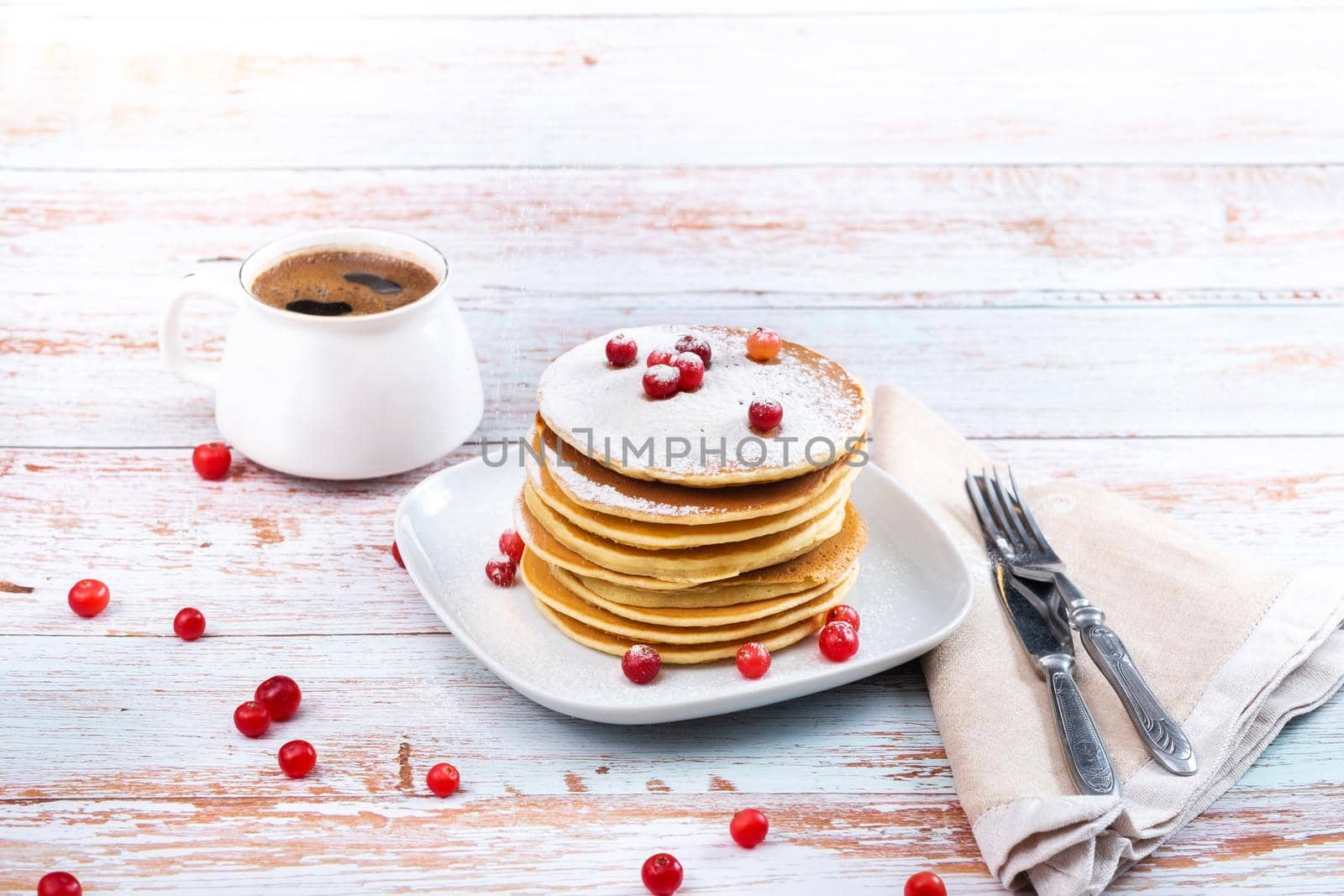 morning breakfast of pancakes with cranberries and powdered sugar on a wooden table and a cup of coffee.