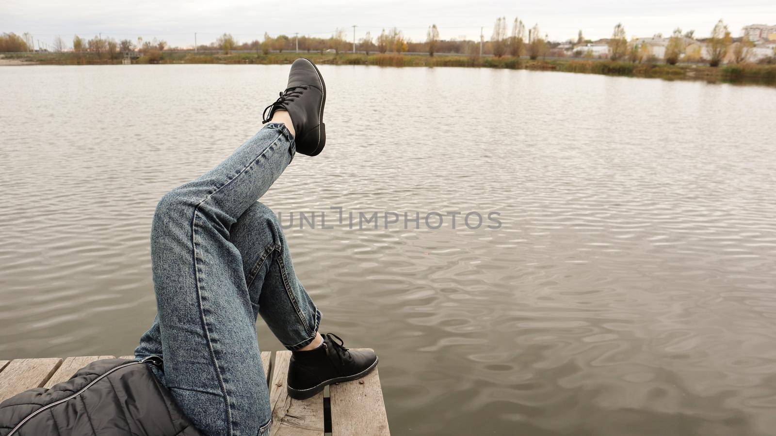 A girl in blue jeans sits by the lake. A young woman is resting by the river, sitting on the edge of a wooden bridge. Pair of legs relaxing by a lake on a wooden dock. People are resting.