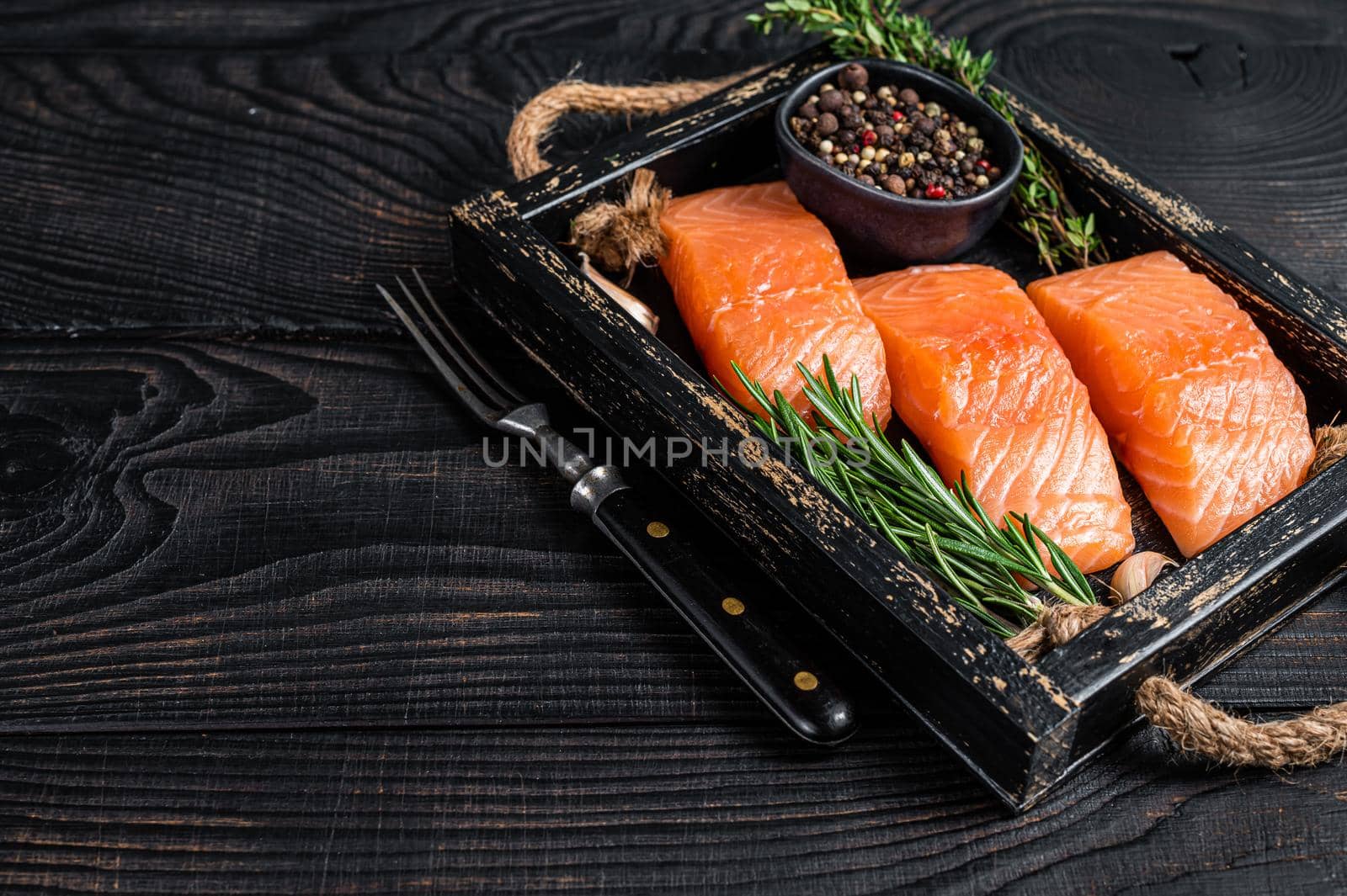 Raw salmon fillet fish steaks in a wooden tray with thyme and rosemary. Black wooden background. Top view. Copy space by Composter