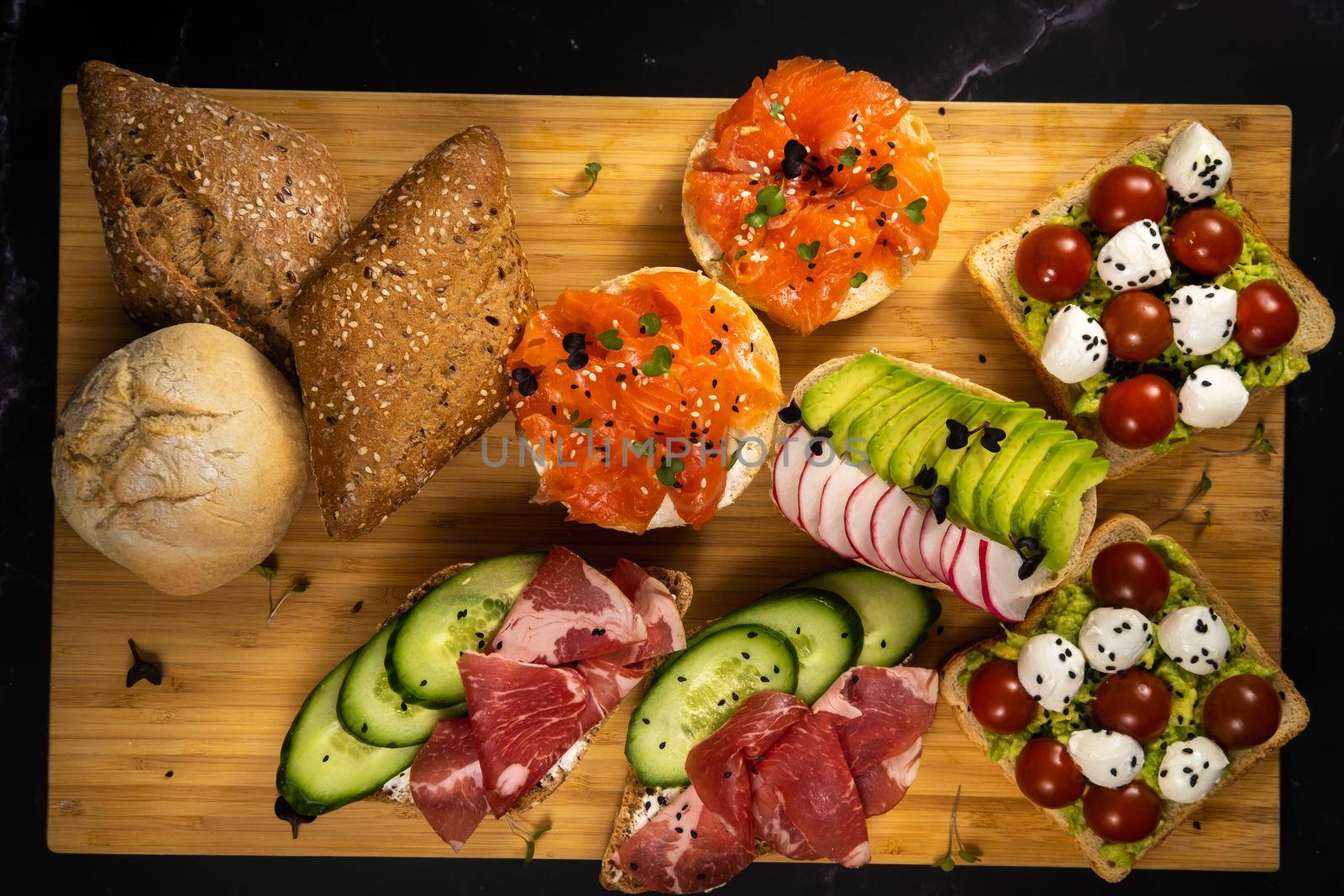 An assortment of sandwiches with fish, cheese, meat and vegetables lay on the board and a bun by Lobachad