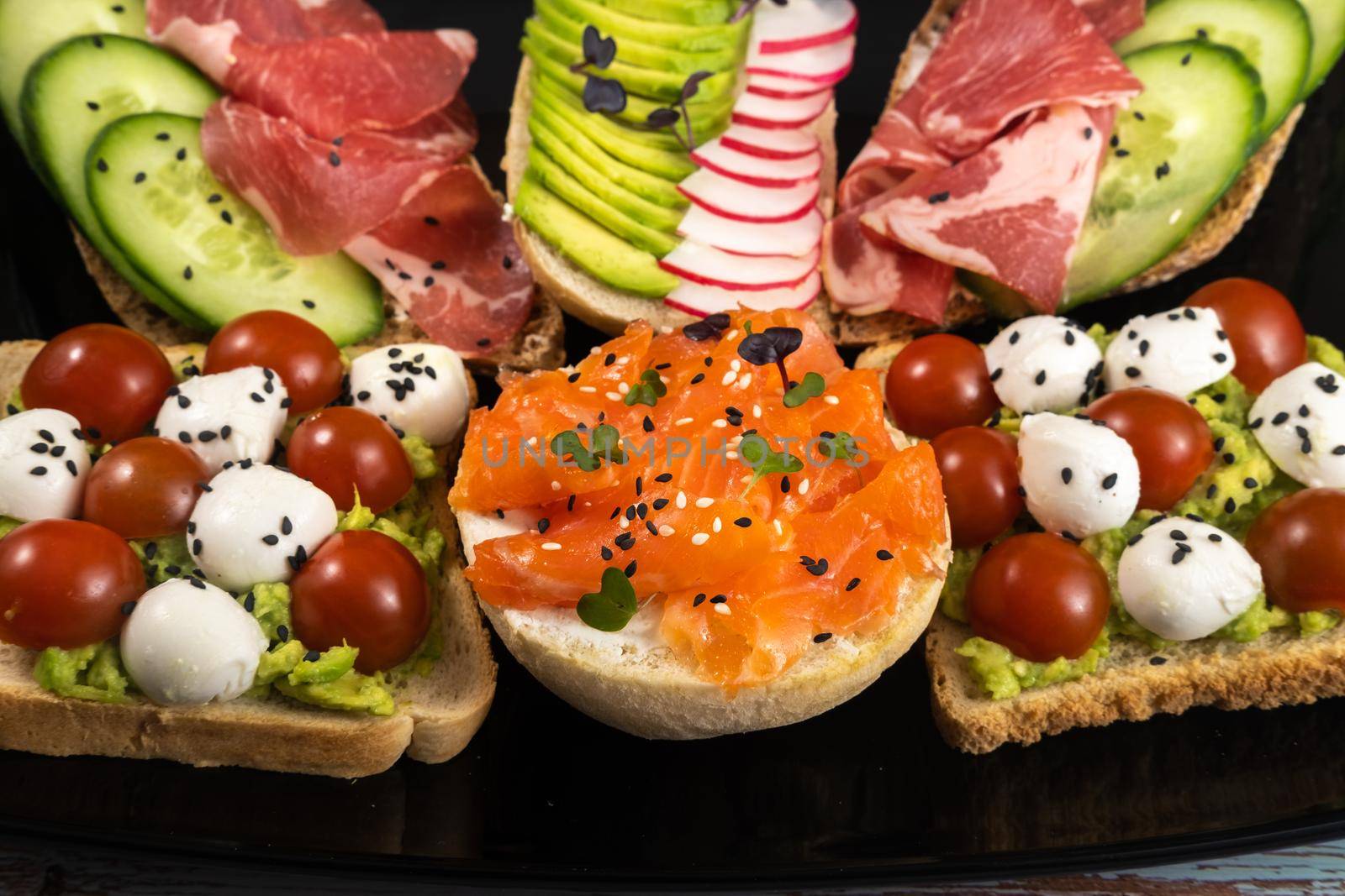 Assorted sandwiches with fish, cheese, meat and vegetables on a black plate and wooden background.