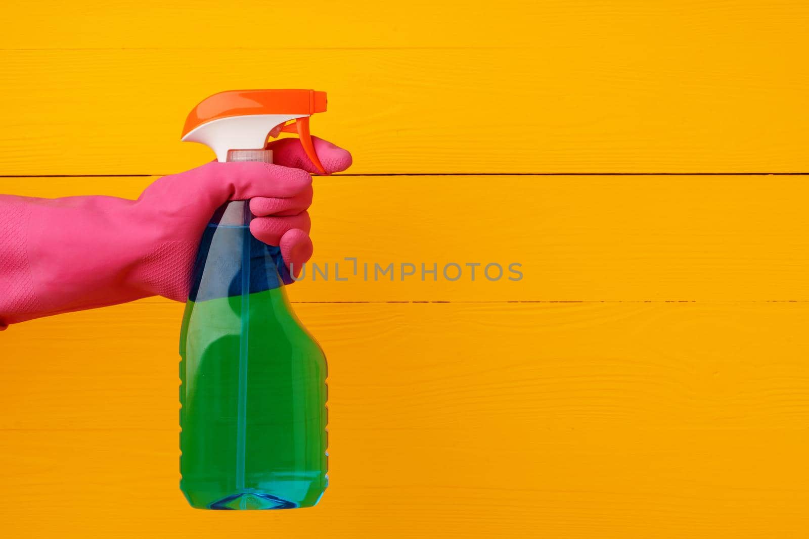 Hand in glove holding household detergent spray against yellow wooden background, close up