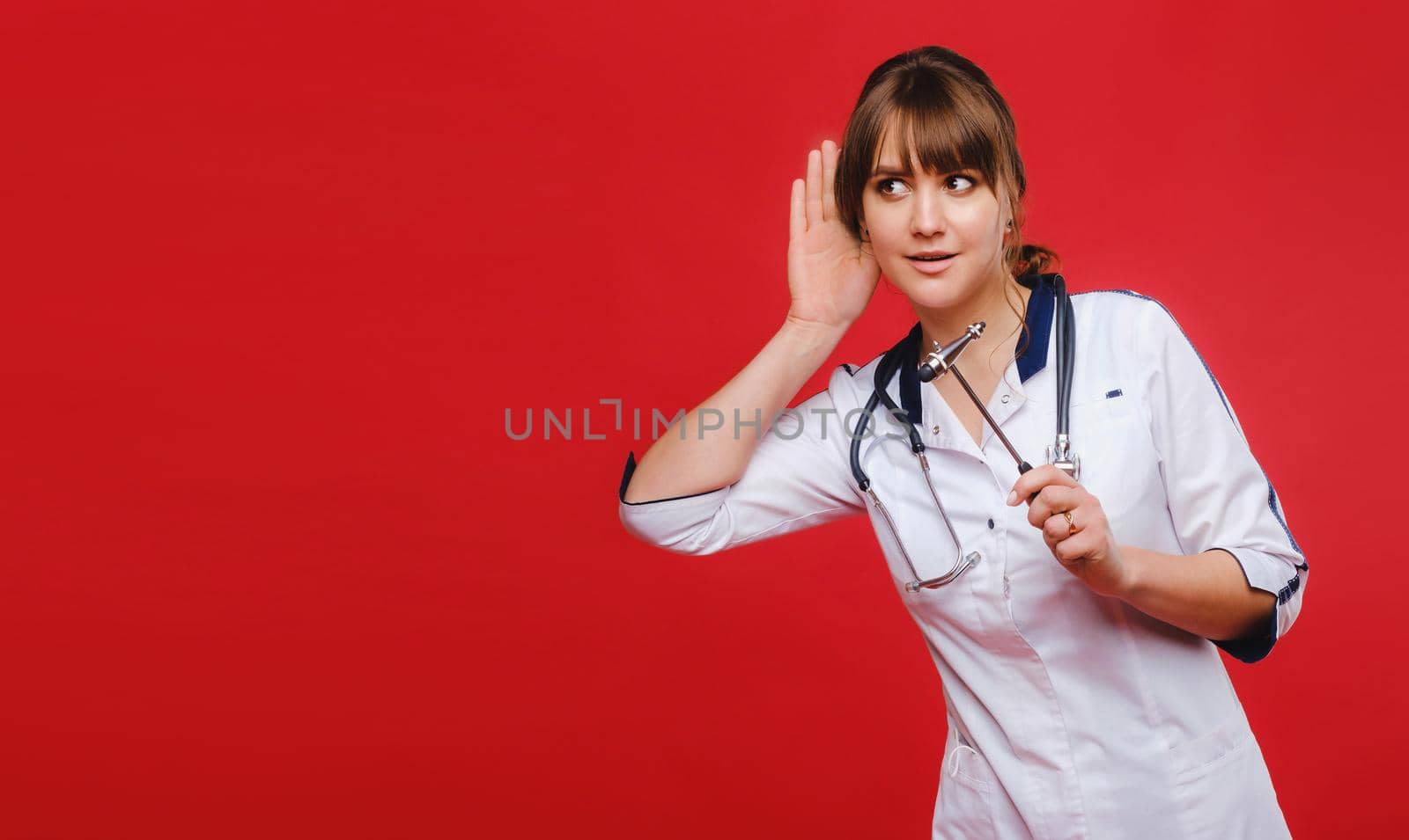 A doctor in a white coat on a red background holds a neurological hammer and listens to something.