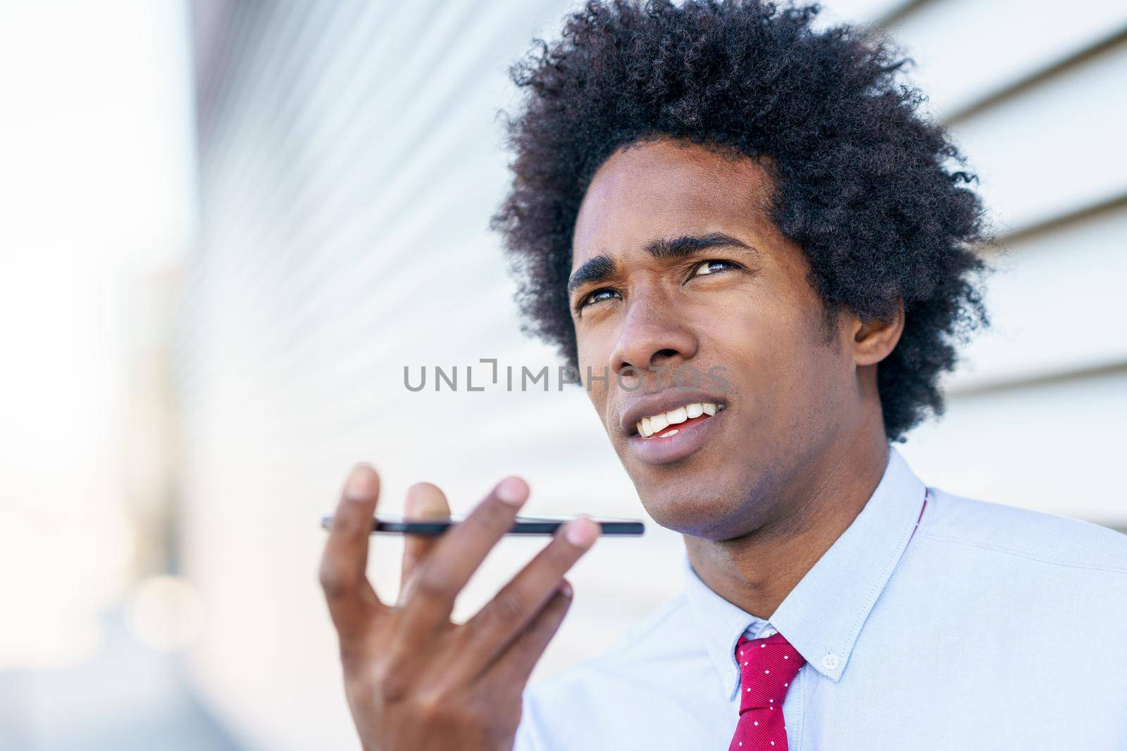 Black Businessman sending voice note with his smartphone near an office building. Man with afro hair.