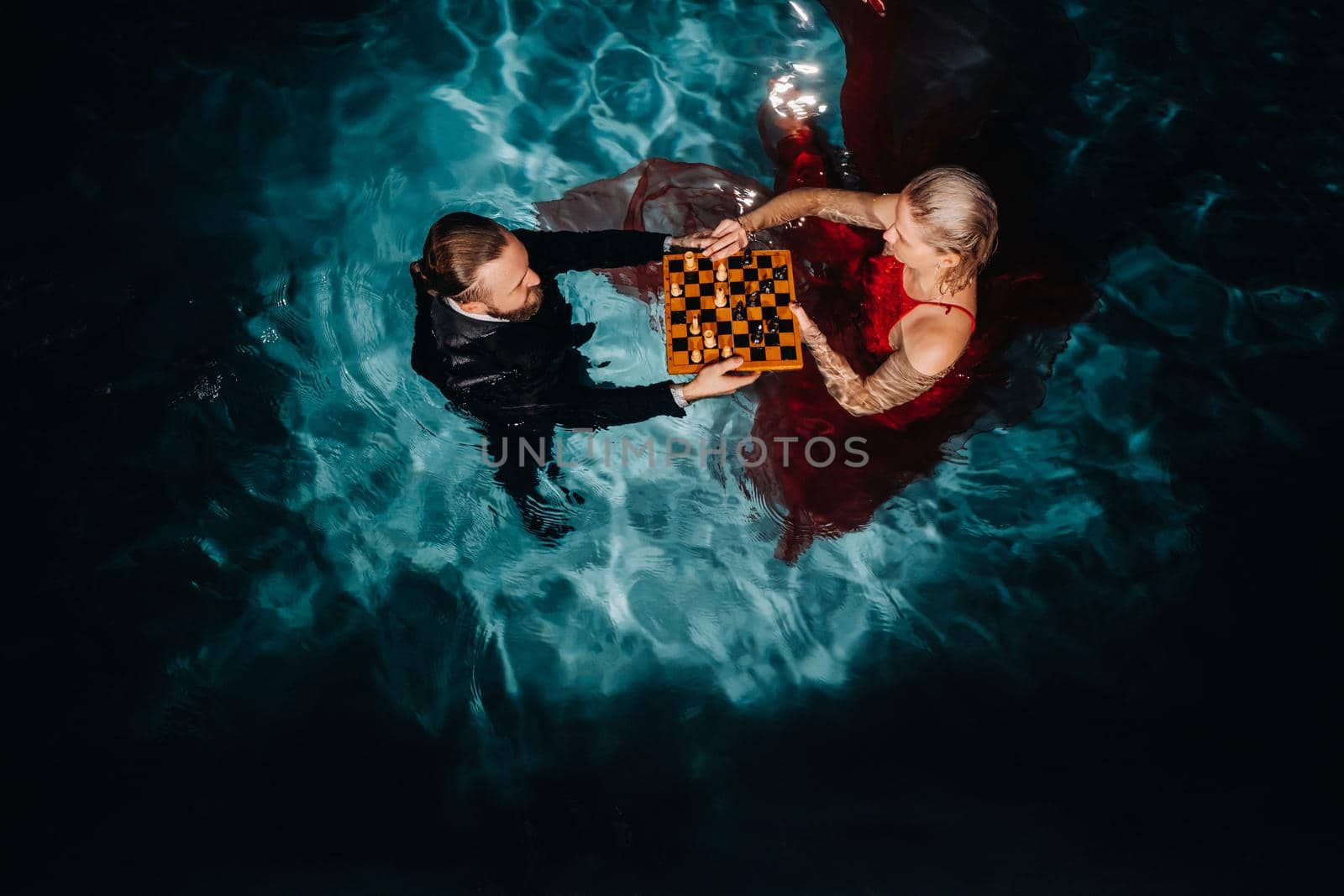 a man in a suit and a girl in a red dress play chess on the water in the pool.