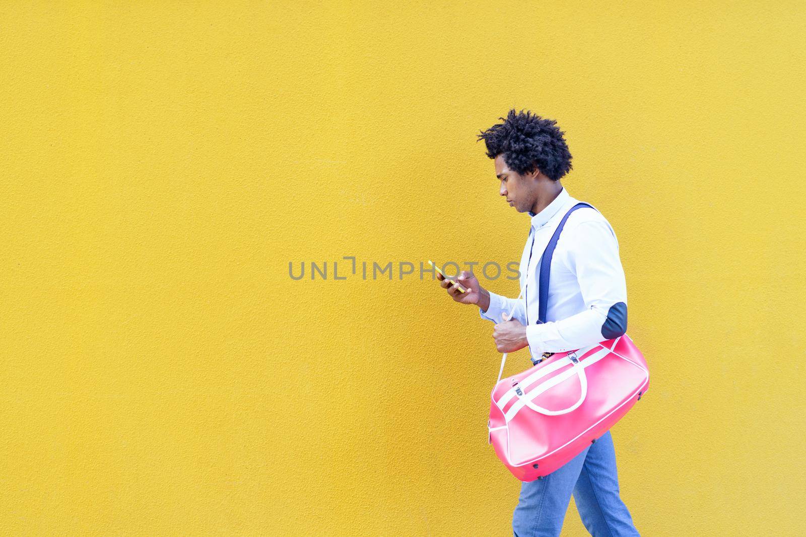 Black man with afro hairstyle carrying a sports bag and smartphone against a yellow urban background. Guy with curly hair wearing shirt and suspenders.