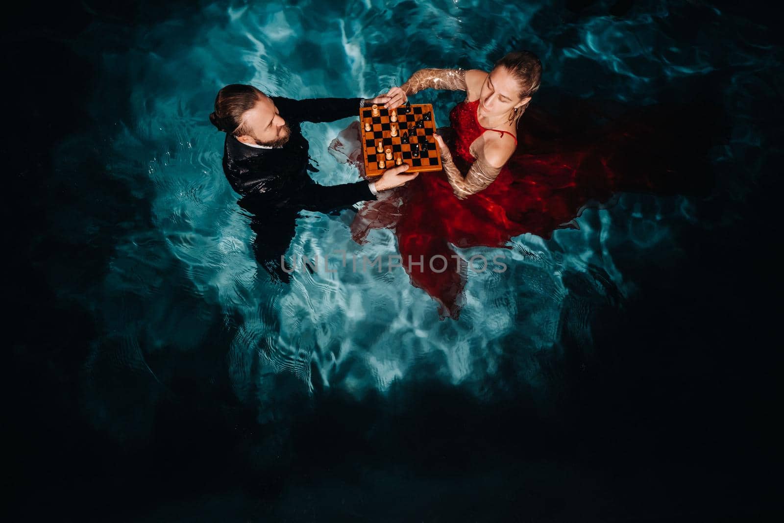 a man in a suit and a girl in a red dress play chess on the water in the pool.