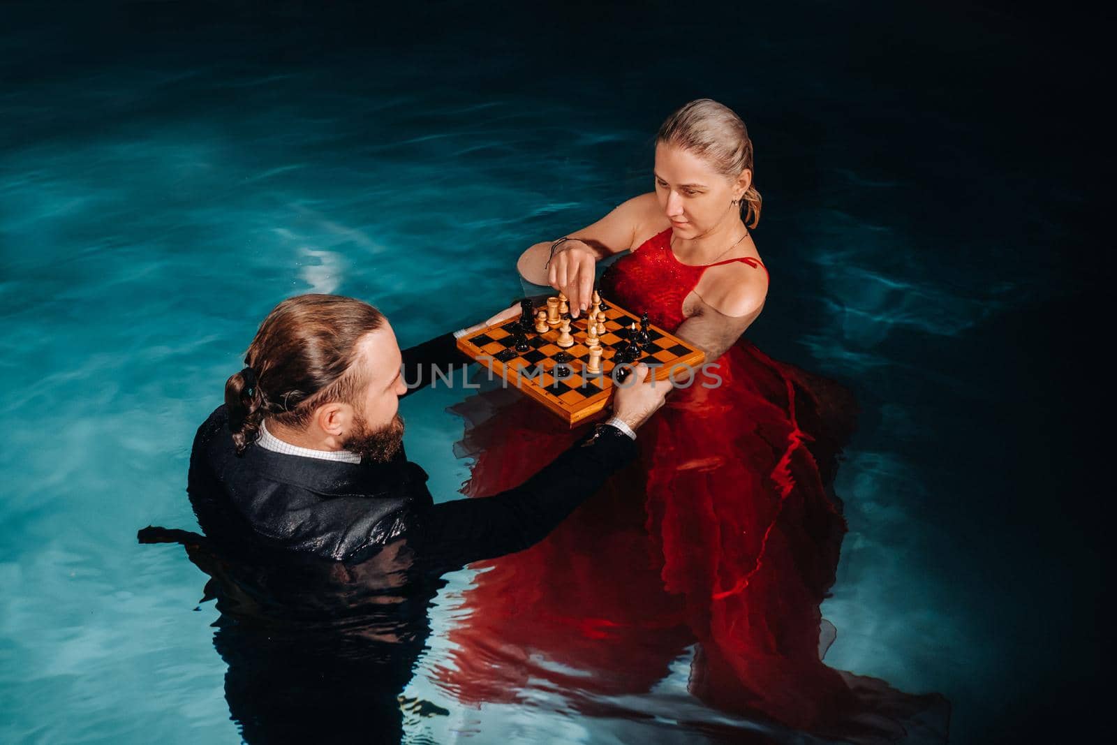 a man in a suit and a girl in a red dress play chess on the water in the pool by Lobachad