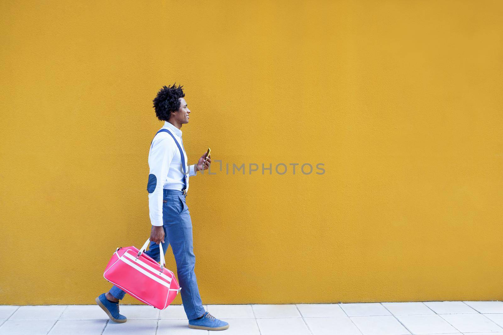 Black man with afro hairstyle carrying a sports bag and smartphone against a yellow urban background. Guy with curly hair wearing shirt and suspenders.