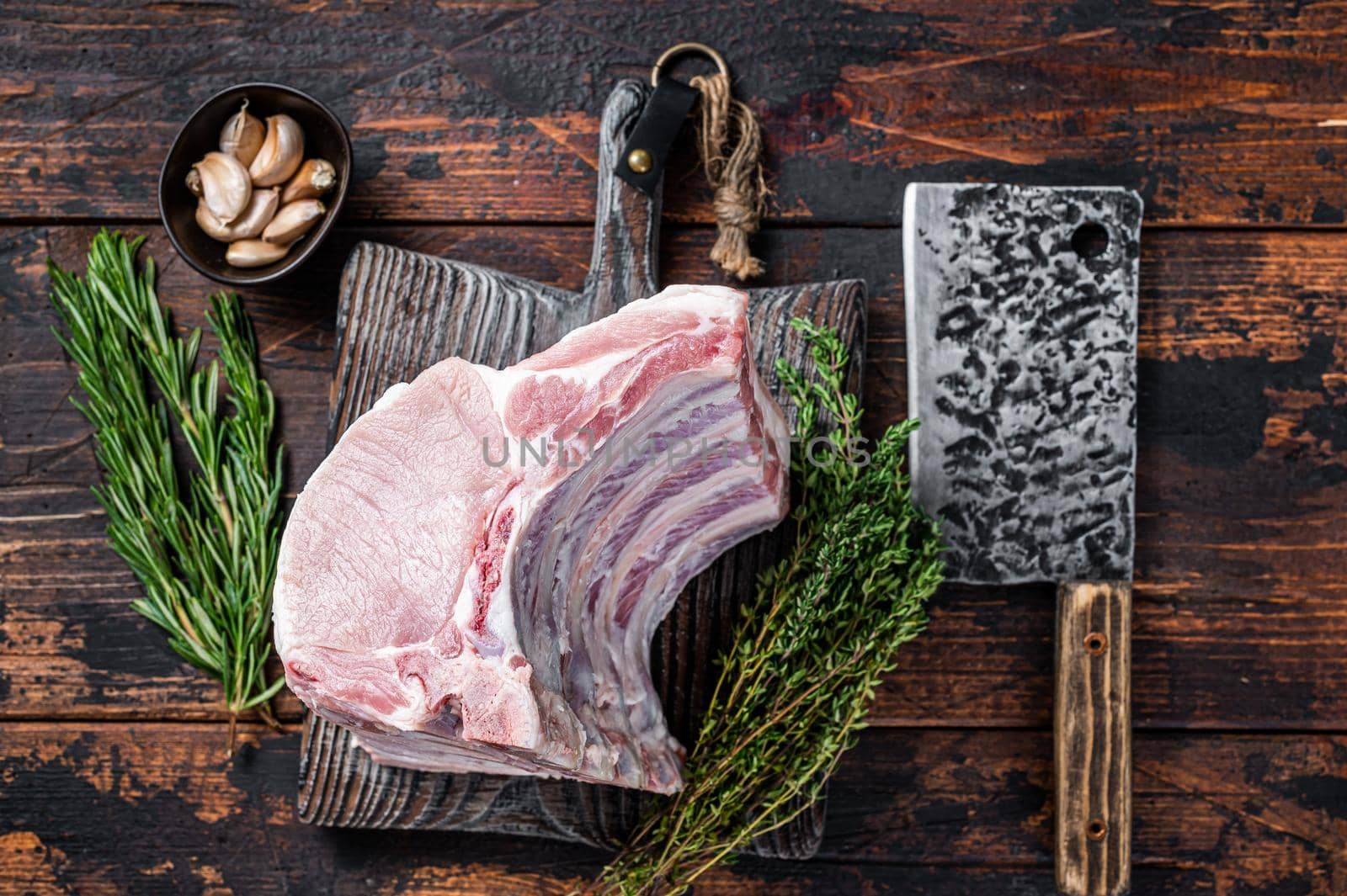 Raw rack of pork loin chops with ribs on a butcher board with meat cleaver. Dark wooden background. Top view.