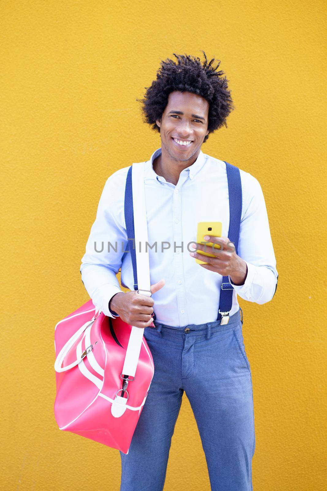 Black man with afro hairstyle carrying a sports bag and smartphone in yellow background. by javiindy