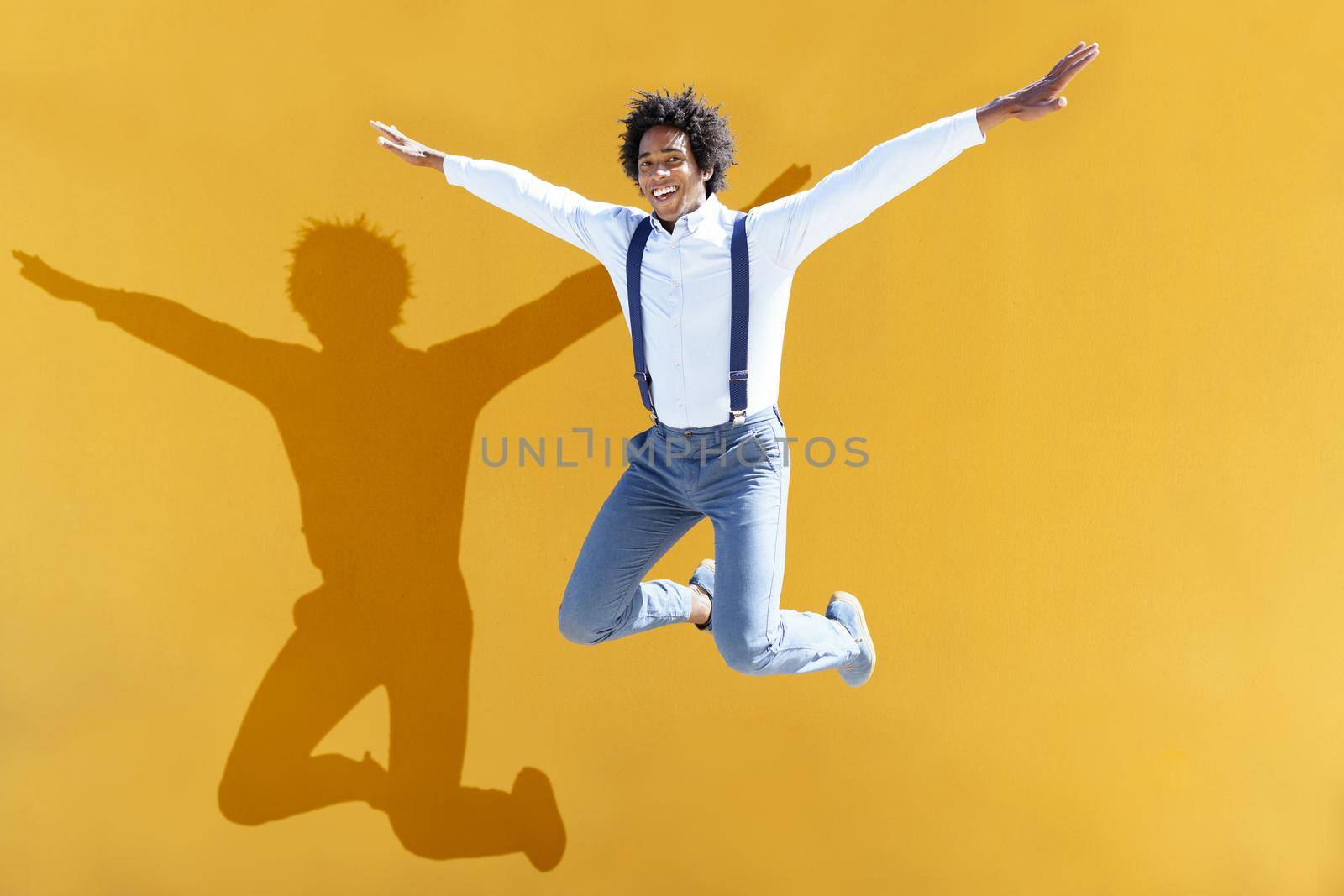 Black man with afro hair jumping on a yellow urban background. Guy wearing shirt and suspenders.