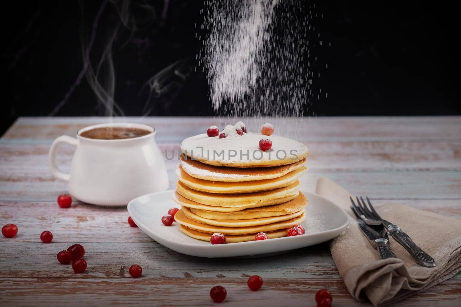 morning breakfast of pancakes with cranberries and powdered sugar on a wooden table and a cup of coffee.