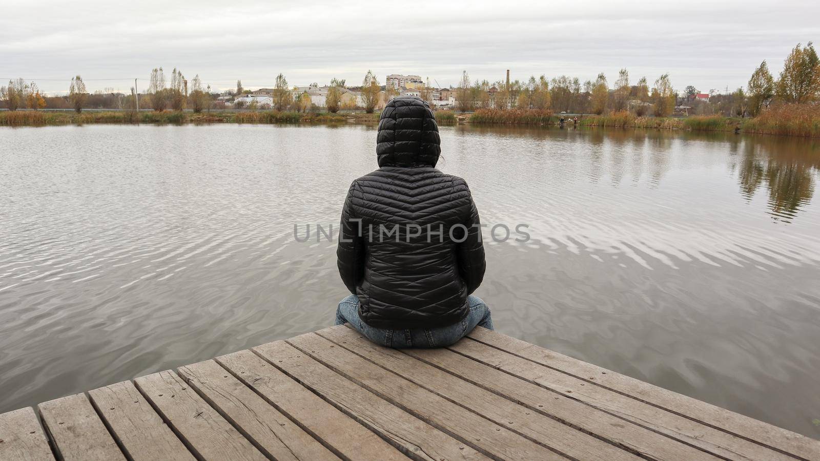 A girl in a jacket sits on a wooden bridge by the lake in cold autumn weather. Dipresia or dreaming alone. Thinking one on one with nature