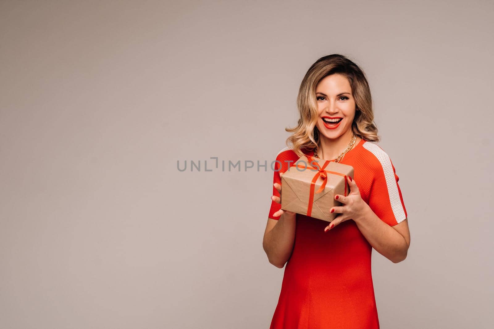 A girl in a red dress stands with gifts in her hands on a gray background.